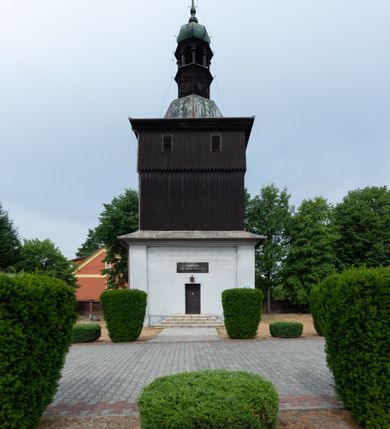 Zdjęcie nr 2: Dzwonnica na planie kwadratu do połowy wysokości murowana, wyżej drewniana, zwieńczona baniastym hełmem z latarnią. 
Część murowana nieco szersza. Od strony wschodniej znajdują się drzwi wejściowe, naroża są wzmocnione lizenami, od północy i południa znajdują się okna prostokątne zamknięte łukiem półokrągłym. Kondygnacje oddziela szeroki fartuch. Górna partia z pozorną izbicą przeprutą z każdej strony dwoma niewielkimi oknami w kształcie stojącego prostokąta zamkniętego łukiem pełnym. Od strony południowej otwór drzwiowy prostokątny zamknięty łukiem półokrągłym. Hełm baniasty przechodzi w oktogon,  na którym są arkadki wspierające mniejszy hełm cebulasty zwieńczony krzyżem. Dachy kryte blachą. 
