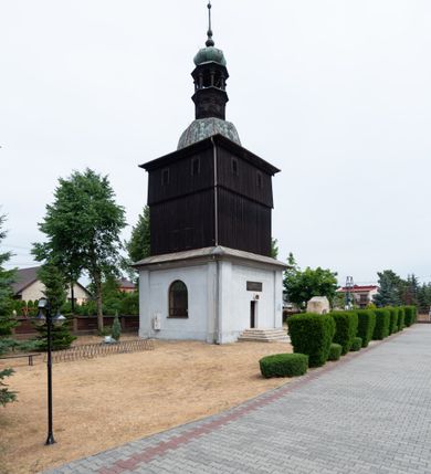 Zdjęcie nr 7: Dzwonnica na planie kwadratu do połowy wysokości murowana, wyżej drewniana, zwieńczona baniastym hełmem z latarnią. 
Część murowana nieco szersza. Od strony wschodniej znajdują się drzwi wejściowe, naroża są wzmocnione lizenami, od północy i południa znajdują się okna prostokątne zamknięte łukiem półokrągłym. Kondygnacje oddziela szeroki fartuch. Górna partia z pozorną izbicą przeprutą z każdej strony dwoma niewielkimi oknami w kształcie stojącego prostokąta zamkniętego łukiem pełnym. Od strony południowej otwór drzwiowy prostokątny zamknięty łukiem półokrągłym. Hełm baniasty przechodzi w oktogon,  na którym są arkadki wspierające mniejszy hełm cebulasty zwieńczony krzyżem. Dachy kryte blachą. 
