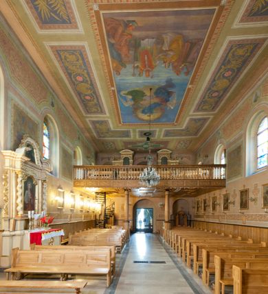 Zdjęcie nr 26: Kościół murowany, tynkowany, orientowany, jednonawowy. Prezbiterium prostokątne, krótkie, zamknięte trójbocznie, przy północnej ścianie prostokątna zakrystia z niższym przedsionkiem. Od wschodu, na zewnętrznej ścianie prezbiterium ogrójec. Nawa główna szersza od prezbiterium, na rzucie prostokąta, od północy przylega prostokątna kaplica, do której od zachodu dobudowany jest niski przedsionek, od południa przylega kruchta boczna, od zachodu prostokątny przedsionek z wieżą na planie kwadratu. Prezbiterium i nawa nakryte stropami, łuk tęczowy półkolisty, wewnątrz ujęty pilastrami, zakończony kluczem. Dostęp do kaplicy z nawy głównej przez wysokie przejście arkadowe na ścianie północnej, w południowej przejście do kruchty bocznej, na osi, pod chórem wejście do przedsionka. Kaplica boczna sklepiona kolebkowo. Fasada bez podziałów z wbudowaną wieżą, ujętą spłaszczonymi spływami. Na osi portal wejściowy flankowany dwoma pilastrami podtrzymującymi gzyms, na którym znajduje się półkoliste okno. W polu dwuskrzydłowe drzwi wejściowe ujęte opaską tynkową. Wieża dwukondygnacyjna, w dolnej kondygnacji okulus, w górnej okna w formie stojącego prostokąta zamkniętego łukiem półkolistym usytuowane z czterech stron. Na spływach ujmujących wieżę dwa okulusy. Wieża nakryta dachem ostrosłupowym. Południowa ściana oraz prezbiterium opięte grubymi szkarpami nakrytymi daszkami o nieregularnej wysokości. Do wschodniej ściany prezbiterium dobudowany murowany ogrojec flankowany filarami, nakryty wielospadowym daszkiem. Elewacje gładkie, tynkowane zakończone profilowanym gzymsem. Wejścia do kościoła przez przybudówkę kaplicy bocznej od północy oraz przez zachodni przedsionek i południową kruchtę. Okna w formie stojącego prostokąta zamkniętego łukiem półkolistym: siedem w elewacji południowej, trzy w północnej. Okna w formie stojącego prostokąta w dwóch kondygnacjach na północnej i południowej ścianie przedsionka zachodniego. Nawa główna, prezbiterium, kaplica i kruchta boczna nakryte dachem dwuspadowym, zakrystia i przybudówki dachami pulpitowymi. 
