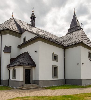 Zdjęcie nr 14: Kościół murowany, tynkowany, orientowany, jednonawowy. Prezbiterium prostokątne, krótkie, zamknięte trójbocznie, przy północnej ścianie prostokątna zakrystia z niższym przedsionkiem. Od wschodu, na zewnętrznej ścianie prezbiterium ogrójec. Nawa główna szersza od prezbiterium, na rzucie prostokąta, od północy przylega prostokątna kaplica, do której od zachodu dobudowany jest niski przedsionek, od południa przylega kruchta boczna, od zachodu prostokątny przedsionek z wieżą na planie kwadratu. Prezbiterium i nawa nakryte stropami, łuk tęczowy półkolisty, wewnątrz ujęty pilastrami, zakończony kluczem. Dostęp do kaplicy z nawy głównej przez wysokie przejście arkadowe na ścianie północnej, w południowej przejście do kruchty bocznej, na osi, pod chórem wejście do przedsionka. Kaplica boczna sklepiona kolebkowo. Fasada bez podziałów z wbudowaną wieżą, ujętą spłaszczonymi spływami. Na osi portal wejściowy flankowany dwoma pilastrami podtrzymującymi gzyms, na którym znajduje się półkoliste okno. W polu dwuskrzydłowe drzwi wejściowe ujęte opaską tynkową. Wieża dwukondygnacyjna, w dolnej kondygnacji okulus, w górnej okna w formie stojącego prostokąta zamkniętego łukiem półkolistym usytuowane z czterech stron. Na spływach ujmujących wieżę dwa okulusy. Wieża nakryta dachem ostrosłupowym. Południowa ściana oraz prezbiterium opięte grubymi szkarpami nakrytymi daszkami o nieregularnej wysokości. Do wschodniej ściany prezbiterium dobudowany murowany ogrojec flankowany filarami, nakryty wielospadowym daszkiem. Elewacje gładkie, tynkowane zakończone profilowanym gzymsem. Wejścia do kościoła przez przybudówkę kaplicy bocznej od północy oraz przez zachodni przedsionek i południową kruchtę. Okna w formie stojącego prostokąta zamkniętego łukiem półkolistym: siedem w elewacji południowej, trzy w północnej. Okna w formie stojącego prostokąta w dwóch kondygnacjach na północnej i południowej ścianie przedsionka zachodniego. Nawa główna, prezbiterium, kaplica i kruchta boczna nakryte dachem dwuspadowym, zakrystia i przybudówki dachami pulpitowymi. 
