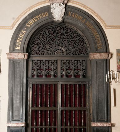 Zdjęcie nr 148: Kaplica zbudowana została na planie kwadratu i nakryta wzniesioną na pendentywach kopułą z latarnią. Przylega do kościoła od północy, do pierwszego przęsła nawy, z którą komunikuje się zamkniętym półkoliście przejściem. Wnętrze artykułowane jest w oparciu o schemat starożytnego łuku triumfalnego za pomocą osadzonych na wysokim cokole pilastrów z kompozytowymi głowicami. Pilastry podtrzymują wydatne belkowanie, ozodbione dekoracją stiukową w postaci puttów, festonów owocowych, rozet oraz kartuszy. Równocześnie pilastry ujmują półkoliste arkady, dwie niższe i większą, środkową. Niższe arkady wypełniają obrazy ze scenami z życia św. Karola Boromeusza. Ponad nimi w prostokątnych, stiukowych, obramieniach znajdują się malowane przedstawienia różnych świętych. W środkowej arkadzie ściany północnej umieszczono okazały późnobarokowy ołtarz z cudownym wizerunkiem św. Karola Boromeusza. Z kolei w arkadach wschodniej i zachodniej rozmieszczono prostokątne okna. Poniżej okna w arkadzie zachodniej znajduje się wyjście na zewnątrz kaplicy zaopatrzone w metalowe, prostokątne drzwi. Natomiast pod oknem w ścianie wschodniej zawieszono tablicę pamiątkową ku czci papieża Jana Pawła II. Arkadę wejściową do kaplicy zamyka prostokątna, brązowa krata. W jej podłuczu od wnętrza na wzór tympanonu umieszczono stiukowy kartusz z herbem Szreniawa, podtrzymywany przez dwa anioły. Ściany tarczowe podniebia przepruto czworobocznymi oknami ujętymi w stiukowe obramienia wolutowe. Na pendentywach przedstawiono w czterech stiukowych kartuszach herby: Szreniawa, Gryf i Kotwicz. Czwarty kartusz jest pusty. Czaszę kopuły wypełnia iluzjonistycznie malowana architektura podzielona na dwie strefy. Dolną artykułują pary pilastrów ujmujące okulusy oraz tralkowa balustrada, a górną gurty wyznaczające pola dla dekoracji ornamentalnej. Ściany zewnętrzne kaplicy zostały podzielone za pomocą kamiennych gzymsów i lizen na dwie trójosiowe kondygnacje, z których górna odpowiada wysokością smukłemu cokołowi mieszczącemu wejście. Zewnętrzne pola każdej elewacji dekorowane są płaskimi pasami tynku w układzie wzoru geometrycznego, zbliżonego do okuciowego. Okna i portal zostały ujęte w profilowane kamienne obramienia. Na ścianie północnej w środkowej kondygnacji pod kartuszem z herbem Szreniawa umieszczono tablicę fundacyjną z napisem: STANISLAUS COMES IN WIŚNICZ LUBOMIRSKI / PALATINUS ET GENERALIS CRACOVIEN[SIS] / SCEPUSIEN[SIS], BIALLOCERKIEVIEN[SIS] / ZATHORIEN[SIS], NIEPOLOMICIEN[SIS] E[T]C[ETER]A, E[T]C[ETER]A, CAPITANEUS / CAPELLAM HANC, / S[ANCTO] CAROLO BARRHOMAEO SUO TUTELARI PATRONO / POST RECEPTA SA[N]CTI HUIUS MERITIS TOT BENEFICIA / DICATAM / EX FUNDAMENTIS EREXIT, DONAVIT / EXORNAVIT, / ANNO D[O]MI[NI], 1640 (Stanisław hrabia na Wiśniczu Lubomirski wojewoda i starosta generalny krakowski, starosta spiski, białocerkiewski, zatorski, niepołomicki, i tak dalej, i tak dalej, tę kaplicę, poświęconą św. Karolowi Boromeuszowi, swojemu patronowi opiekuńczemu, po otrzymaniu tak wielkich łask za wstawiennictwem tego świetego, od fundamentów wzniósł, uposażył i ozdobił, roku Pańskiego 1640). Dach kaplicy oraz latarnia pokryte są blachą miedzianą.
