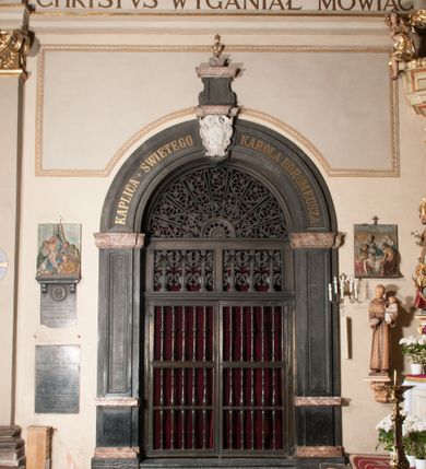 Zdjęcie nr 149: Kaplica zbudowana została na planie kwadratu i nakryta wzniesioną na pendentywach kopułą z latarnią. Przylega do kościoła od północy, do pierwszego przęsła nawy, z którą komunikuje się zamkniętym półkoliście przejściem. Wnętrze artykułowane jest w oparciu o schemat starożytnego łuku triumfalnego za pomocą osadzonych na wysokim cokole pilastrów z kompozytowymi głowicami. Pilastry podtrzymują wydatne belkowanie, ozodbione dekoracją stiukową w postaci puttów, festonów owocowych, rozet oraz kartuszy. Równocześnie pilastry ujmują półkoliste arkady, dwie niższe i większą, środkową. Niższe arkady wypełniają obrazy ze scenami z życia św. Karola Boromeusza. Ponad nimi w prostokątnych, stiukowych, obramieniach znajdują się malowane przedstawienia różnych świętych. W środkowej arkadzie ściany północnej umieszczono okazały późnobarokowy ołtarz z cudownym wizerunkiem św. Karola Boromeusza. Z kolei w arkadach wschodniej i zachodniej rozmieszczono prostokątne okna. Poniżej okna w arkadzie zachodniej znajduje się wyjście na zewnątrz kaplicy zaopatrzone w metalowe, prostokątne drzwi. Natomiast pod oknem w ścianie wschodniej zawieszono tablicę pamiątkową ku czci papieża Jana Pawła II. Arkadę wejściową do kaplicy zamyka prostokątna, brązowa krata. W jej podłuczu od wnętrza na wzór tympanonu umieszczono stiukowy kartusz z herbem Szreniawa, podtrzymywany przez dwa anioły. Ściany tarczowe podniebia przepruto czworobocznymi oknami ujętymi w stiukowe obramienia wolutowe. Na pendentywach przedstawiono w czterech stiukowych kartuszach herby: Szreniawa, Gryf i Kotwicz. Czwarty kartusz jest pusty. Czaszę kopuły wypełnia iluzjonistycznie malowana architektura podzielona na dwie strefy. Dolną artykułują pary pilastrów ujmujące okulusy oraz tralkowa balustrada, a górną gurty wyznaczające pola dla dekoracji ornamentalnej. Ściany zewnętrzne kaplicy zostały podzielone za pomocą kamiennych gzymsów i lizen na dwie trójosiowe kondygnacje, z których górna odpowiada wysokością smukłemu cokołowi mieszczącemu wejście. Zewnętrzne pola każdej elewacji dekorowane są płaskimi pasami tynku w układzie wzoru geometrycznego, zbliżonego do okuciowego. Okna i portal zostały ujęte w profilowane kamienne obramienia. Na ścianie północnej w środkowej kondygnacji pod kartuszem z herbem Szreniawa umieszczono tablicę fundacyjną z napisem: STANISLAUS COMES IN WIŚNICZ LUBOMIRSKI / PALATINUS ET GENERALIS CRACOVIEN[SIS] / SCEPUSIEN[SIS], BIALLOCERKIEVIEN[SIS] / ZATHORIEN[SIS], NIEPOLOMICIEN[SIS] E[T]C[ETER]A, E[T]C[ETER]A, CAPITANEUS / CAPELLAM HANC, / S[ANCTO] CAROLO BARRHOMAEO SUO TUTELARI PATRONO / POST RECEPTA SA[N]CTI HUIUS MERITIS TOT BENEFICIA / DICATAM / EX FUNDAMENTIS EREXIT, DONAVIT / EXORNAVIT, / ANNO D[O]MI[NI], 1640 (Stanisław hrabia na Wiśniczu Lubomirski wojewoda i starosta generalny krakowski, starosta spiski, białocerkiewski, zatorski, niepołomicki, i tak dalej, i tak dalej, tę kaplicę, poświęconą św. Karolowi Boromeuszowi, swojemu patronowi opiekuńczemu, po otrzymaniu tak wielkich łask za wstawiennictwem tego świetego, od fundamentów wzniósł, uposażył i ozdobił, roku Pańskiego 1640). Dach kaplicy oraz latarnia pokryte są blachą miedzianą.
