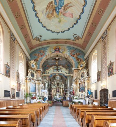 Zdjęcie nr 23: Kościół murowany, tynkowany, jednonawowy. Prezbiterium prostokątne, zamknięte prosto, z obu stron przy nawie prostokątne, piętrowe lokalności (zakrystia i skarbiec, empory). Nawa szersza od prezbiterium, na rzucie prostokąta, od frontu przylega wieża na planie kwadratu z dwoma aneksami po bokach. Prezbiterium dwuprzęsłowe, podzielone wewnątrz pilastrami, sklepione żagielkowo na gurtach. Empory flankowane parami półkolumienek, z oknami w formie leżącego prostokąta zamkniętego łukiem odcinkowym, nad lewą herb Odrowąż z inicjałami „JG”. Nawa nakryta pozornym, spłaszczonym sklepieniem kolebkowym. Otwór tęczowy w formie półkolistej arkady wspartej na pilastrach, z belką o fantazyjnym kształcie. Chór muzyczny dwukondygnacyjny, wsparty na dwóch filarach i dwóch półfilarach, pomiędzy którymi arkady ostrołukowe, kondygnacja zamknięta profilowanym gzymsem. Druga kondygnacja z trzema arkadami, w środkowej organ z wklęsło-wypukłą, ażurową balustradą. Posadzka z płyt ceramicznych. 
Fasada trójosiowa, dwukondygnacyjna. Pierwsza kondygnacja wydzielona pilastrami dekorowanymi blendami, zwieńczona profilowanym gzymsem z fryzem arkadkowym. Nad pilastrami osi bocznych dwie stylizowane wieżyczki, zakończone krzyżami. 
Na każdej osi ostrołukowe arkady, w środkowej wejście, dwie boczne wypełnione stylizowanymi blendami; nad nimi otwory okienne. Na osi środkowej, w drugiej kondygnacji wieża, ujęta w narożach czterema wieżyczkami, ścianki flankowane lizenami, zakończona profilowanym gzymsem z kostkowaniem. Na ściankach z czterech stron tarcze zegarowe oraz otwory dzwonne w formie ostrołukowych arkad w opaskach tynkowych. Wieża nakryta dachem ostrosłupowym. 
Elewacje gładkie, tynkowane zakończone profilowanym gzymsem. Wyodrębnione aneksy elewacji bocznych ujęte grubymi lizenami, z ostrołukowymi arkadami w polu, ujętymi opaskami tynkowymi. Od strony fasady w partiach aneksów bocznych wejścia do kościoła, od strony prezbiterium ostrołukowe okienka. Na każdej elewacji bocznej po trzy okna w formie stojącego prostokąta zamkniętego łukiem półkolistym, o szerokich rozglifieniach, po dwa w nawie i jednym w prezbiterium. Elewacja zamykająca prezbiterium trójosiowa, wydzielona lizenami, z głębokimi wnękami w formie stojącego prostokąta zamkniętego łukiem półkolistym, z namalowanym wizerunkiem Ukrzyżowanego Chrystusa i dwóch łotrów na krzyżach. Elewacja zakończona profilowanym gzymsem, z daszkiem pulpitowym oraz trójkątnym szczytem z czterema cokołami. Nawa i prezbiterium nakryte osobnymi  dachami dwuspadowymi. Na połączeniu nawy i aneksów przy prezbiterium wieżyczka na sygnaturkę z latarnią, nakryta dachem ostrosłupowym. 


