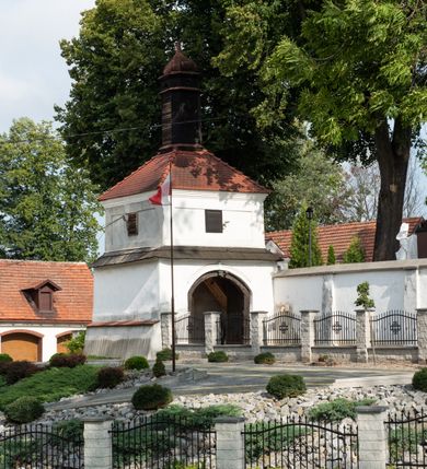 Zdjęcie nr 12: Kościół jest usytuowany w centrum wsi Rudawa, na północ od głównej drogi z Zabierzowa do Chrzanowa. Teren wokół niego jest otoczony wysokim, kamiennym murem z nieregularnych ciosów kamienia, oszkarpowanym, tynkowanym, z wyjątkiem zewnętrznej części muru na zachód od południowej bramki. W murze znajdują się trzy wejścia, od wschodu, południa i zachodu. W części wschodniej muru znajduje się trójosiowa brama wjazdowa, zwieńczona łukiem wklęsło-wypukłym, z trzema przelotami: środkowym wyższym i szerszym, zamkniętym łukiem odcinkowym i bocznymi węższymi i niższymi, zamkniętymi łukiem półkolistym (północne zamurowane). Część środkowa jest flankowana zdwojonymi pilastrami, podtrzymującymi profilowane uskokowe belkowanie z przyczółkiem o profilowanym łuku falistym, zwieńczonym równoramiennym krzyżem. W przyczółku wnęka z figurą św. Floriana. Bramki od strony zachodniej i południowej prostokątne, z przejściem zakończonym płaskim łukiem odcinkowym, z profilowanym gzymsem, zwieńczonym trójkątnym przyczółkiem. Przejścia zamykane drewnianymi ażurowymi i pomalowanymi na ciemnobrązowo jednoskrzydłowymi drzwiami. Poprzez bramkę od zachodu zejście kamiennymi schodami poprowadzonymi wzdłuż muru na dziedziniec plebański, usytuowany poniżej terenu kościelnego. 
W północno-wschodnim narożniku muru niewielka murowana i tynkowana kostnica na planie kwadratu, nakryta dachem namiotowym, łamanym, zakończonym ośmioboczną ślepą latarnią.

Po stronie południowej, poniżej placu kościelnego, do muru przylega murowana, tynkowana, prostokątna i dwukondygnacyjna „zegarowa” wieża bramna nakryta namiotowym dachem z wysoką drewnianą ośmioboczną latarnią o cebulastym hełmie. W wieży znajduje się przejazd wiodący na dziedziniec plebański, z sienią otwartą na przestrzał w dolnej kondygnacji i z lamusem dostępnym od strony placu kościelnego w górnej. 

W południowej części muru, w pobliżu południowo-wschodniego narożnika placu kościelnego, nadbudowana kamienna dzwonnica parawanowa z zawieszonymi czterema dzwonami.  

We wschodniej części placu kościelnego pomiędzy bramką wschodnią a kostnicą znajduje się kamienna ambona, o ośmiobocznej podstawie przechodzącej w szerszy, również ośmioboczny korpus, zakończony kamiennym parapetem, z kamiennymi schodami prowadzącymi na ambonę od strony wschodniej. 

Kościół jest orientowany, prawie w całości zbudowany z cegły, otynkowany z wyjątkiem prezbiterium. Wnętrze kościoła tworzy niewielkie, prostokątne, jednoprzęsłowe prezbiterium zakończone prostą ścianą i nakryte pozornym sklepieniem krzyżowo-żebrowym. Nawa jest szersza, prawie kwadratowa, nakryta drewnianym stropem. Prezbiterium z nawą są połączone szeroką, ostrołukową arkadą w ścianie tęczowej. Od północy do prezbiterium przylega założona na planie prostokąta dwuprzęsłowa zakrystia, nakryta sklepieniem krzyżowym. Wejście do zakrystii zaznaczone gotyckim, kamiennym, sfazowanym portalem zamkniętym łukiem ostrym. Od zachodu do zakrystii przylega niewielkie pomieszczenie gospodarcze, do którego z zakrystii prowadzą ostrołukowe drzwi. Zakrystia oświetlona ostrołukowym oknem w ścianie wschodniej, wypełnionym witrażem z tafelek w kształcie sześciokąta foremnego, ze szkła surowego, tzw. katedralnego. 

Od południa do nawy przylega kaplica o formach renesansowych, dwukondygnacyjna, założona na planie kwadratu. W dolnej kondygnacji sklepiona krzyżowo-żebrowo kruchta otwarta do nawy szeroką, zamkniętą półkoliście arkadą.

Ponad kruchtą kaplica św. Kazimierza, nakryta wspartą na pendentywach kopułą z latarnią. Z kruchty do kaplicy prowadzą kręte kamienne schody umieszczone od zachodu w półkolistej wieżyczce. Prostokątne wejście z krypty na klatkę schodową ujęte kamiennym profilowanym portalem. Kaplica otwarta do nawy szeroką, zamkniętą półkoliście arkadą, o kształcie i wymiarach analogicznych do arkady znajdującej się poniżej. Kaplicę, prócz latarni, oświetla prostokątne okno w ścianie południowej, wypełnione witrażem z tafelek w kształcie gomółek, ze szkła surowego, tzw. katedralnego. Na zachodniej ścianie nawy chór muzyczny, dostępny klatką schodową ukrytą w zewnętrznym aneksie przylegającym do zachodniej elewacji nawy i południowej elewacji wieży-dzwonnicy. 

Nawę i prezbiterium oświetlają umieszczone w południowych ścianach prostokątne, rozglifione okna: jedno w prezbiterium, dwa w nawie. Wszystkie wypełnione witrażami z tafelek w kształcie gomółek, ze szkła surowego, tzw. katedralnego. 

Do kościoła prowadzą dwa wejścia: poprzez arkadę z kruchty południowej oraz szerokie wejście główne prostokątne, zamknięte łukiem odcinkowym, prowadzące do kościoła z obszernej kruchty od strony zachodniej, znajdującej się w murowanym przyziemiu wieży - dzwonnicy. Kruchta, założona na planie kwadratu i nakryta murowanym stropem, doświetlona jest dwoma prostokątnymi oknami umieszczonymi naprzeciw siebie w ścianach północnej i południowej.
 
W zewnętrznej elewacji kościoła widoczny niewysoki kamienny cokół wykonany z nieregularnych ciosów kamienia. Prezbiterium, niższe od nawy, murowane z cegły w układzie wedyjskim, z domieszką układu polskiego. Wschodnie naroża prezbiterium zaakcentowane płaskimi lizenami. We wschodniej ścianie widoczny zarys zamurowanego wydłużonego okna zamkniętego lekko zaostrzonym łukiem, w trójkątnym szczycie znajduje się niewielkie wąskie okienko zakończone łukiem półkolistym. Południowa ściana prezbiterium ozdobiona fryzem z cegieł. Do ściany północnej prezbiterium przylega zakrystia, oszkarpowana, nakryta dachem pulpitowym, z dobudowanym od niej niewielkim aneksem, nakrytym takim samym zadaszeniem. Do wschodniej ściany prezbiterium przylega ogrójec nakryty dachem namiotowym z cebulastym zwieńczeniem, wspartym na sześciu smukłych, profilowanych drewnianych słupach z mieczami, pomiędzy którymi od północy i południa ażurowa drewniana balustrada. 

Nawa oszkarpowana w narożnikach. Elewacje bez podziałów architektonicznych. Od strony południowej do nawy dobudowana dwukondygnacyjna kaplica nakryta kopułą z latarnią, podzielona na dwie kondygnacje. W dolnej kondygnacji od południa wejście do kruchty zaznaczone kamiennym portalem o sfazowanych węgarach i lekko zaostrzonym łuku. Wejście zamykają drewniane dwuskrzydłowe drzwi ozdobione kratownicą z żelaznych listew. Od zachodu do kaplicy przylega półokrągła wieżyczka nakryta daszkiem stożkowym. 

Do zachodniej fasady kościoła dostawiona niska, założona na planie kwadratu trójkondygnacyjna wieża-dzwonnica: w dolnej części murowana, w narożach wzmocniona kamiennymi szkarpami, w górnej drewniana konstrukcji słupowo-ramowej z nadwieszoną izbicą i nakryta dachem półszczytowym. W dolnej, murowanej kondygnacji, na osi kościoła, prostokątne wejście do kruchty zaakcentowane przedsionkiem w kształcie ostrołukowej arkady i czterostopniowymi schodami. Przedsionek nakryty dachem dwuspadowym. Do zachodniej elewacji nawy i południowej elewacji dzwonnicy przylega niewielki, wysoki drewniany aneks, nakryty dachem pulpitowym, mieszczący klatkę schodową prowadzącą na górną kondygnację dzwonnicy i na chór muzyczny w nawie kościoła. Prostokątne wejście w południowej ścianie aneksu, do którego prowadzą pięciostopniowe proste schody. Ponad nimi, na wysokości drugiej kondygnacji dzwonnicy, niewielkie kwadratowe okno. 

Prezbiterium i nawa przykryte stromymi dachami dwuspadowymi, zamkniętymi od wschodu i zachodu szczytami sterczynowymi. Wieżyczka na sygnaturkę ośmioboczna, w dolnej części barokowa, w górnej części nowsza.