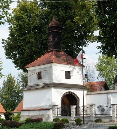 Zdjęcie nr 18: Kościół jest usytuowany w centrum wsi Rudawa, na północ od głównej drogi z Zabierzowa do Chrzanowa. Teren wokół niego jest otoczony wysokim, kamiennym murem z nieregularnych ciosów kamienia, oszkarpowanym, tynkowanym, z wyjątkiem zewnętrznej części muru na zachód od południowej bramki. W murze znajdują się trzy wejścia, od wschodu, południa i zachodu. W części wschodniej muru znajduje się trójosiowa brama wjazdowa, zwieńczona łukiem wklęsło-wypukłym, z trzema przelotami: środkowym wyższym i szerszym, zamkniętym łukiem odcinkowym i bocznymi węższymi i niższymi, zamkniętymi łukiem półkolistym (północne zamurowane). Część środkowa jest flankowana zdwojonymi pilastrami, podtrzymującymi profilowane uskokowe belkowanie z przyczółkiem o profilowanym łuku falistym, zwieńczonym równoramiennym krzyżem. W przyczółku wnęka z figurą św. Floriana. Bramki od strony zachodniej i południowej prostokątne, z przejściem zakończonym płaskim łukiem odcinkowym, z profilowanym gzymsem, zwieńczonym trójkątnym przyczółkiem. Przejścia zamykane drewnianymi ażurowymi i pomalowanymi na ciemnobrązowo jednoskrzydłowymi drzwiami. Poprzez bramkę od zachodu zejście kamiennymi schodami poprowadzonymi wzdłuż muru na dziedziniec plebański, usytuowany poniżej terenu kościelnego. 
W północno-wschodnim narożniku muru niewielka murowana i tynkowana kostnica na planie kwadratu, nakryta dachem namiotowym, łamanym, zakończonym ośmioboczną ślepą latarnią.

Po stronie południowej, poniżej placu kościelnego, do muru przylega murowana, tynkowana, prostokątna i dwukondygnacyjna „zegarowa” wieża bramna nakryta namiotowym dachem z wysoką drewnianą ośmioboczną latarnią o cebulastym hełmie. W wieży znajduje się przejazd wiodący na dziedziniec plebański, z sienią otwartą na przestrzał w dolnej kondygnacji i z lamusem dostępnym od strony placu kościelnego w górnej. 

W południowej części muru, w pobliżu południowo-wschodniego narożnika placu kościelnego, nadbudowana kamienna dzwonnica parawanowa z zawieszonymi czterema dzwonami.  

We wschodniej części placu kościelnego pomiędzy bramką wschodnią a kostnicą znajduje się kamienna ambona, o ośmiobocznej podstawie przechodzącej w szerszy, również ośmioboczny korpus, zakończony kamiennym parapetem, z kamiennymi schodami prowadzącymi na ambonę od strony wschodniej. 

Kościół jest orientowany, prawie w całości zbudowany z cegły, otynkowany z wyjątkiem prezbiterium. Wnętrze kościoła tworzy niewielkie, prostokątne, jednoprzęsłowe prezbiterium zakończone prostą ścianą i nakryte pozornym sklepieniem krzyżowo-żebrowym. Nawa jest szersza, prawie kwadratowa, nakryta drewnianym stropem. Prezbiterium z nawą są połączone szeroką, ostrołukową arkadą w ścianie tęczowej. Od północy do prezbiterium przylega założona na planie prostokąta dwuprzęsłowa zakrystia, nakryta sklepieniem krzyżowym. Wejście do zakrystii zaznaczone gotyckim, kamiennym, sfazowanym portalem zamkniętym łukiem ostrym. Od zachodu do zakrystii przylega niewielkie pomieszczenie gospodarcze, do którego z zakrystii prowadzą ostrołukowe drzwi. Zakrystia oświetlona ostrołukowym oknem w ścianie wschodniej, wypełnionym witrażem z tafelek w kształcie sześciokąta foremnego, ze szkła surowego, tzw. katedralnego. 

Od południa do nawy przylega kaplica o formach renesansowych, dwukondygnacyjna, założona na planie kwadratu. W dolnej kondygnacji sklepiona krzyżowo-żebrowo kruchta otwarta do nawy szeroką, zamkniętą półkoliście arkadą.

Ponad kruchtą kaplica św. Kazimierza, nakryta wspartą na pendentywach kopułą z latarnią. Z kruchty do kaplicy prowadzą kręte kamienne schody umieszczone od zachodu w półkolistej wieżyczce. Prostokątne wejście z krypty na klatkę schodową ujęte kamiennym profilowanym portalem. Kaplica otwarta do nawy szeroką, zamkniętą półkoliście arkadą, o kształcie i wymiarach analogicznych do arkady znajdującej się poniżej. Kaplicę, prócz latarni, oświetla prostokątne okno w ścianie południowej, wypełnione witrażem z tafelek w kształcie gomółek, ze szkła surowego, tzw. katedralnego. Na zachodniej ścianie nawy chór muzyczny, dostępny klatką schodową ukrytą w zewnętrznym aneksie przylegającym do zachodniej elewacji nawy i południowej elewacji wieży-dzwonnicy. 

Nawę i prezbiterium oświetlają umieszczone w południowych ścianach prostokątne, rozglifione okna: jedno w prezbiterium, dwa w nawie. Wszystkie wypełnione witrażami z tafelek w kształcie gomółek, ze szkła surowego, tzw. katedralnego. 

Do kościoła prowadzą dwa wejścia: poprzez arkadę z kruchty południowej oraz szerokie wejście główne prostokątne, zamknięte łukiem odcinkowym, prowadzące do kościoła z obszernej kruchty od strony zachodniej, znajdującej się w murowanym przyziemiu wieży - dzwonnicy. Kruchta, założona na planie kwadratu i nakryta murowanym stropem, doświetlona jest dwoma prostokątnymi oknami umieszczonymi naprzeciw siebie w ścianach północnej i południowej.
 
W zewnętrznej elewacji kościoła widoczny niewysoki kamienny cokół wykonany z nieregularnych ciosów kamienia. Prezbiterium, niższe od nawy, murowane z cegły w układzie wedyjskim, z domieszką układu polskiego. Wschodnie naroża prezbiterium zaakcentowane płaskimi lizenami. We wschodniej ścianie widoczny zarys zamurowanego wydłużonego okna zamkniętego lekko zaostrzonym łukiem, w trójkątnym szczycie znajduje się niewielkie wąskie okienko zakończone łukiem półkolistym. Południowa ściana prezbiterium ozdobiona fryzem z cegieł. Do ściany północnej prezbiterium przylega zakrystia, oszkarpowana, nakryta dachem pulpitowym, z dobudowanym od niej niewielkim aneksem, nakrytym takim samym zadaszeniem. Do wschodniej ściany prezbiterium przylega ogrójec nakryty dachem namiotowym z cebulastym zwieńczeniem, wspartym na sześciu smukłych, profilowanych drewnianych słupach z mieczami, pomiędzy którymi od północy i południa ażurowa drewniana balustrada. 

Nawa oszkarpowana w narożnikach. Elewacje bez podziałów architektonicznych. Od strony południowej do nawy dobudowana dwukondygnacyjna kaplica nakryta kopułą z latarnią, podzielona na dwie kondygnacje. W dolnej kondygnacji od południa wejście do kruchty zaznaczone kamiennym portalem o sfazowanych węgarach i lekko zaostrzonym łuku. Wejście zamykają drewniane dwuskrzydłowe drzwi ozdobione kratownicą z żelaznych listew. Od zachodu do kaplicy przylega półokrągła wieżyczka nakryta daszkiem stożkowym. 

Do zachodniej fasady kościoła dostawiona niska, założona na planie kwadratu trójkondygnacyjna wieża-dzwonnica: w dolnej części murowana, w narożach wzmocniona kamiennymi szkarpami, w górnej drewniana konstrukcji słupowo-ramowej z nadwieszoną izbicą i nakryta dachem półszczytowym. W dolnej, murowanej kondygnacji, na osi kościoła, prostokątne wejście do kruchty zaakcentowane przedsionkiem w kształcie ostrołukowej arkady i czterostopniowymi schodami. Przedsionek nakryty dachem dwuspadowym. Do zachodniej elewacji nawy i południowej elewacji dzwonnicy przylega niewielki, wysoki drewniany aneks, nakryty dachem pulpitowym, mieszczący klatkę schodową prowadzącą na górną kondygnację dzwonnicy i na chór muzyczny w nawie kościoła. Prostokątne wejście w południowej ścianie aneksu, do którego prowadzą pięciostopniowe proste schody. Ponad nimi, na wysokości drugiej kondygnacji dzwonnicy, niewielkie kwadratowe okno. 

Prezbiterium i nawa przykryte stromymi dachami dwuspadowymi, zamkniętymi od wschodu i zachodu szczytami sterczynowymi. Wieżyczka na sygnaturkę ośmioboczna, w dolnej części barokowa, w górnej części nowsza.