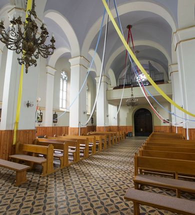 Zdjęcie nr 24: Kościół orientowany, murowany, trójnawowy, czteroprzęsłowy, z jednoprzęsłowym prezbiterium zakończonym wielobocznie. Do prezbiterium dostawione symetrycznie aneksy, od zachodu kruchta, w przyziemiu wieża flankowana dwiema klatkami schodowymi (pomieszczeniem gospodarczym?). Nawy oddzielone od siebie masywnymi filarami opilastrowanymi, z uproszczonymi kapitelami, podtrzymującymi arkady. Nawy i prezbiterium nakryte sklepieniem krzyżowym na gurtach, pięcioboczne zamknięcie prezbiterium sklepieniem parasolowatym. Nawę i prezbiterium oświetlają wysokie, prostokątne okna zakończone półkoliście z oknami trójrzędowymi dwuosiowymi, zakończonymi maswerkami. Układ komunikacyjny zaznaczony kamiennymi portalami o sfazowanych węgarach. Do kościoła prowadzą duże prostokątne drzwi, w kruchcie zakończone półkoliście. Tam też prostokątne wejścia do aneksów bocznych. W południowo-zachodniej ścianie prostokątne wejście zamknięte ściętym łukiem nadwieszonym. Takie samo wejście do aneksu południowego przy prezbiterium. Do aneksu północnego przy prezbiterium prostokątne wejście zamknięte łukiem odcinkowym, ujęte kamiennym portalem, zamkniętym półkoliście. Wejście w ścianie nawy południowej zaznaczone kamiennym portalem zamkniętym łukiem ostrym. W skrajnym przęśle nawy od zachodu chór muzyczny wsparty na dwóch filarach międzynawowych. W części chóru nad kruchtą wnęka otwarta kamienną rozetą. Fasada kościoła trójosiowa, dwukondygnacyjna, ujęta szkarpami z uskokami. Szkarpy z uskokami wydzielają także środkową oś fasady. W jej dolnej kondygnacji znajduje się kamienny portal główny, ujęty uskokowo ustawionymi półkolumnami i zakończony półkolistym tympanonem wypełnionym płaskorzeźbą ze sceną Zesłania Ducha Świętego. Powyżej wypełniona maswerkiem rozeta w profilowanej ramie. Między nią a biforium niewielki herb rodziny Potockich. Szczyt dekoruje fryz arkadkowy. W osiach bocznych wysokie, dwudzielne okna z maswerkiem. Budynek ustawiony na kamiennym cokole, oszkarpowany, z kamiennymi detalami, dekorowany fryzem arkadkowym, z otwartym od zewnątrz obejściem prezbiterium. W obejściu płaskorzeźbiona grupa Ukrzyżowania. Dachy kryte blachą – nawy i wieży dwuspadowe, przybudówek przy prezbiterium pulpitowe, nad obejściem i prezbiterium wieloboczne. Na kalenicy wieżyczka na sygnaturkę z latarnią i wielobocznym dachem. 