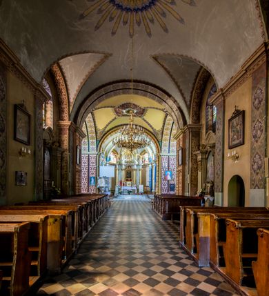 Zdjęcie nr 24: Kościół murowany, jednonawowy, z wieżą częściowo wtopioną w elewację zachodnią składający się z nawy, transeptu, zakrystii od północy z mieszczącym się na piętrze skarbcem i przystawionej od wschodu wieżyczki klatki schodowej oraz kruchty od południa. Prezbiterium dwuprzęsłowe, zamknięte trójbocznie. Nawa główna podzielona masywnymi opilastrowanymi filarami. Sklepienie prezbiterium i nawy kolebkowe z lunetami, skrzyżowania z transeptem. Od południa chór muzyczny mieszczący się między pierwszymi filarami i powyżej kruchty wejściowej. W prezbiterium okna w każdym przęśle, po każdej stronie nawy po dwa, w transepcie po trzy, w kruchcie dwa. Główne wejście od zachodu, kolejne do zakrystii i od południa. Ściany i sklepienia kościoła pokrywa polichromia złożona z geometryzujących dekoracji floralnych. 
Wschodnią elewację frontową kościoła tworzy trójosiowa, wieżowa fasada. Oś kompozycji stanowi wysunięta ryzalitowo dwukondygnacyjna wieża ze ściętymi narożami, które zaakcentowane są poprzez pilastry. Ściana czołowa wieży wypełniona w pierwszej kondygnacji wklęsłą płyciną, z portalem o wykroju prostokątnym, z odcinkiem gzymsu w zwieńczeniu. Ponad nim prostokątne okno zakończone łukiem pełnym. Kondygnacje rozdzielone za pomocą masywnego gzymsu. Druga kondygnacja wieży wypełniona analogicznie prostokątną płyciną, w którą wkomponowany jest wypukły panel w kształcie leżącego prostokąta. Nad nim okno o obramieniu i wykroju identycznym jak w kondygnacji niższej. Całość zwieńczona za pomocą gzymsu podokapowego i baniastego hełmu wieżowego o ściętych narożach, na które nałożona jest kopułka w formie cebulastej. Osie boczne cofnięte w stosunku do wysuniętej wieży i zrytmizowane podziałem ramowym. Tak jak w osi głównej pierwsza kondygnacja wypełniona jest wklęsłą płyciną w formie wydłużonego, stojącego prostokąta. Nad gzymsem trójkątny spływ przysłonięty wieżą. Elewacje boczne i tylna ukształtowane jednorodnie. Zrytmizowane podziałem ramowym i przeprute oknami umieszczonymi w prostokątnych płycinach. Ramiona transeptu oraz prezbiterium zrytmizowane są za pomocą pilastrów załamanych na narożach. Od południa do korpusu przylega niska kruchta. Prowadzi do niej wejście o wykroju prostokątnym z profilowanym w tynku portalem i z odcinkiem gzymsu w zwieńczeniu. Od północy pomiędzy prezbiterium, a ramieniem transeptu wprowadzona została piętrowa przybudówka zakrystii. W elewacji tej partii kościoła brak jest znaczących podziałów. Okna i drzwi tam się znajdujące mają formę stojącego prostokąta i pozbawione są jakichkolwiek obramień. Kościół pokryty dwuspadowym, jednokalenicowym dachem, nad zakrystią i kruchtami umieszczono daszki dwuspadowe. Wieża nakryta jest hełmem cebulastym. Całość pokryta została malowaną blachą.
