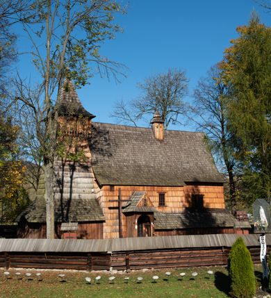 Zdjęcie nr 21: Orientowany, jednoprzestrzenny, nawa na rzucie prostokąta, z węższym, prostokątnym prezbiterium, przy którym od północy wąska zakrystia. Od zachodu przy nawie wieża na rzucie kwadratu, od południa kruchta. Wnętrze nakryte stropem, w nawie z zaskrzynieniem. Portale zachodni i do zakrystii ostrołukowe, fazowane; portal południowy zamknięty łukiem kotarowym. Okna prostokątne, w prezbiterium od południa okno bliźnie, od wschodu zamknięte łukiem kotarowym.  Wokół podcienia, obecnie zaszalowane. Więźba dachowa jętkowa, ze znakami montażowymi.  Dach nad nawą i prezbiterium jednokalenicowy, dwuspadowy, nad nawą z połaciami przedłużonymi na zaskrzynienia i od północy nad zakrystię. Na kalenicy wieżyczka na sygnaturkę z baniastym hełmem.  Wieża o ścianach pochyłych, z nadwieszoną izbicą, zwieńczona ostrosłupowym hełmem. Wokół kościoła ogrodzenie belek układanych na zrąb, z dwiema bramkami. 