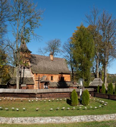 Zdjęcie nr 24: Orientowany, jednoprzestrzenny, nawa na rzucie prostokąta, z węższym, prostokątnym prezbiterium, przy którym od północy wąska zakrystia. Od zachodu przy nawie wieża na rzucie kwadratu, od południa kruchta. Wnętrze nakryte stropem, w nawie z zaskrzynieniem. Portale zachodni i do zakrystii ostrołukowe, fazowane; portal południowy zamknięty łukiem kotarowym. Okna prostokątne, w prezbiterium od południa okno bliźnie, od wschodu zamknięte łukiem kotarowym.  Wokół podcienia, obecnie zaszalowane. Więźba dachowa jętkowa, ze znakami montażowymi.  Dach nad nawą i prezbiterium jednokalenicowy, dwuspadowy, nad nawą z połaciami przedłużonymi na zaskrzynienia i od północy nad zakrystię. Na kalenicy wieżyczka na sygnaturkę z baniastym hełmem.  Wieża o ścianach pochyłych, z nadwieszoną izbicą, zwieńczona ostrosłupowym hełmem. Wokół kościoła ogrodzenie belek układanych na zrąb, z dwiema bramkami. 