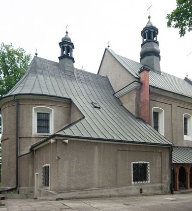 Zdjęcie nr 16: Kościół murowany, orientowany, jednonawowy (z dodatkową przestrzenią od południa powstałą w wyniku połączenia trzech kaplic), z półkoliście zamkniętym prezbiterium, z prostokątną zakrystią od północy, z kruchtą na osi nawy głównej oraz drugą od północy; między zakrystią a kruchtą północną babiniec. 
Nawa szersza od prezbiterium, prostokątna, trójprzęsłowa, ze sklepieniem kolebkowym z lunetami na gurtach. Od zachodu chór muzyczny wsparty na kolumnach. Ściany nawy podzielone zdwojonymi pilastrami, między którymi nisze arkadowe, od południa półokrągłe arkady łączące nawę główną z nawą boczną. Partię ścian od sklepienia oddziela pełne belkowanie wyłamane na osi pilastrów, które jest kontynuowane w przekrytym sklepieniem kolebkowym z lunetami prezbiterium.  
W partii przyziemia oraz sklepienia prostokątne okna w rozglifionych obramieniach zamkniętych łukiem odcinkowym, na osi prezbiterium owalne. Od zachodu oraz od południa drzwi zamknięte łukiem odcinkowym. 
Fasada trójosiowa, dwukondygnacyjna, zwieńczona trójkątnym przyczółkiem, poprzedzona jednokondygnacyjną kruchtą, do której wchodzi się po sześciostopniowych schodach. Osie rozdzielone są pojedynczymi lub zdwojonymi pilastrami, kondygnacje dzieli bogato profilowane belkowanie. W kruchcie drzwi o wykroju prostokąta zamknięte łukiem odcinkowym, okna w osiach bocznych o identycznym jak drzwi wykroju; okulus w szczycie. W osi środkowej drugiej kondygnacji fasady okno prostokątne, zamknięte łukiem odcinkowym, a u szczytu okulus. 
Na elewacjach kontynuowany jest gzyms oddzielający drugą kondygnację i partię dachu, pionowe podziały utworzone zostały przez lizeny. 
Nawa główna oraz kruchty nakryte dachami dwuspadowymi, część nawy bocznej, zakrystia oraz babiniec jednospadowymi, druga część nawy bocznej trójspadowym, zakrystia dwuspadowym zamkniętym półkoliście. Dwie wieżyczki na sygnaturki: jedna nad nawą główną, druga, mniejsza nad prezbiterium. Obie przeprute arkadami, zwieńczone kopułami z kulą i krzyżem. 
Do najważniejszych elementów wyposażenia należą: ołtarz główny późnobarokowy z obrazem Przemienienia Pańskiego przywieziony z Rzymu w 1723 roku, dwa regencyjne ołtarze boczne z barokowymi obrazami Matki Boskiej z Dzieciątkiem oraz św. Katarzyny, oba ozdobione sukienkami oraz obraz z przedstawieniem św. Józefa, pochodzący z trzeciego ołtarza wisi teraz na ścianie nawy głównej. 
