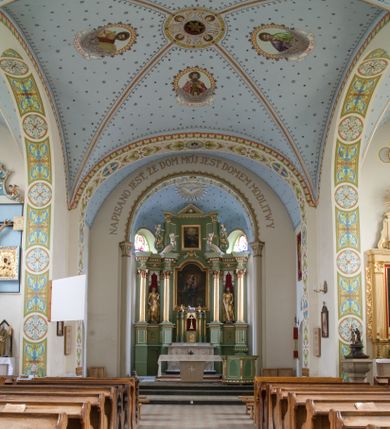 Zdjęcie nr 19: Kościół na planie krzyża łacińskiego, jednonawowy, pięcioprzęsłowy, z transeptem i jednoprzęsłowym prezbiterium zakończonym wielobocznie. Ramiona transeptu jednoprzęsłowe, o wysokości nawy, zamknięte prostymi ścianami. W narożnikach pomiędzy prezbiterium i ramionami transeptu dwa prostokątne aneksy mieszczące zakrystię i skarbiec. Prezbiterium i ramiona transeptu oddzielone od nawy wydatnymi półfilarami przechodzącymi bezpośrednio w gurty sklepienne. Prezbiterium od absydy oddziela arkada tęczowa wsparta na dostawionych do węgarów kolumnach kompozytowych. Sklepienie nad skrzyżowaniem naw krzyżowe, nad pozostałymi przęsłami nawy kolebkowe o przekroju koszowym, nad ramionami transeptu krzyżowe (ostrołukowe), w absydzie ćwierćsferyczne z lunetami. Otwory okienne prostokątne, zamknięte półkoliście, rozglifione do wewnątrz; trzy pary w nawie, dwa na ścianach zamykających ramiona transeptu. Ponad oknami w transepcie dodatkowo po jednym okrągłym oknie wypełnionym rozetą. W prezbiterium okna w kształcie wydłużonego prostokąta o smukłych proporcjach zamkniętego półkoliście, pomiędzy nimi para regularnie rozmieszczonych okien owalnych.  W zachodnim przęśle nawy murowany trójprzęsłowy chór muzyczny wsparty na parze ośmiobocznych filarów z cokołami, a pod ścianami na parze półfilarów. Balustrada o prostej dekoracji płycinowej. Podniebie chóru podzielone belkami na trzy kasetony, pośrodku których kwadratowe płyciny. Fasada kościoła dwukondygnacyjna, trójosiowa przechodząca w czworoboczną wieżę. Ściany rozczłonkowane lizenami, kondygnacje oddzielone profilowanym gzymsem. W części środkowej wejście zamknięte łukiem półkolistym, z dwuspadowym drewnianym daszkiem, wyżej prostokątne okno zamknięte półkoliście. W polach bocznych mniejsze, prostokątne okna zamknięte półkoliście. Wieża o narożach ujętych lizenami, podtrzymującymi gzyms, nakryta ozdobnym zwieńczeniem z czterema wieżyczkami w narożach, pomiędzy którymi trójkątne szczyciki nad oknami i ostrosłupowy hełm zakończony krzyżem. Ściany boczne nawy i transeptu ujęte uskokowymi przyporami. Ściany zamykające ramiona transeptu podzielone na dwie strefy, z których dolna ujęta lizenami, górna wypełniona oknem w ozdobnym obramieniu. Dachy dwuspadowe, kryte dachówką. Na skrzyżowaniu naw wieloboczna, drewniana wieżyczka na sygnaturkę nakryta ostrosłupowym hełmem pokrytym blachą.

