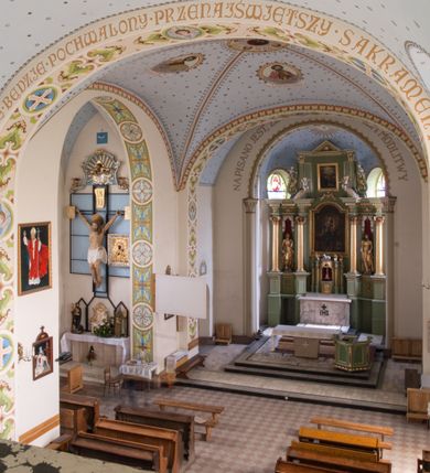 Zdjęcie nr 24: Kościół na planie krzyża łacińskiego, jednonawowy, pięcioprzęsłowy, z transeptem i jednoprzęsłowym prezbiterium zakończonym wielobocznie. Ramiona transeptu jednoprzęsłowe, o wysokości nawy, zamknięte prostymi ścianami. W narożnikach pomiędzy prezbiterium i ramionami transeptu dwa prostokątne aneksy mieszczące zakrystię i skarbiec. Prezbiterium i ramiona transeptu oddzielone od nawy wydatnymi półfilarami przechodzącymi bezpośrednio w gurty sklepienne. Prezbiterium od absydy oddziela arkada tęczowa wsparta na dostawionych do węgarów kolumnach kompozytowych. Sklepienie nad skrzyżowaniem naw krzyżowe, nad pozostałymi przęsłami nawy kolebkowe o przekroju koszowym, nad ramionami transeptu krzyżowe (ostrołukowe), w absydzie ćwierćsferyczne z lunetami. Otwory okienne prostokątne, zamknięte półkoliście, rozglifione do wewnątrz; trzy pary w nawie, dwa na ścianach zamykających ramiona transeptu. Ponad oknami w transepcie dodatkowo po jednym okrągłym oknie wypełnionym rozetą. W prezbiterium okna w kształcie wydłużonego prostokąta o smukłych proporcjach zamkniętego półkoliście, pomiędzy nimi para regularnie rozmieszczonych okien owalnych.  W zachodnim przęśle nawy murowany trójprzęsłowy chór muzyczny wsparty na parze ośmiobocznych filarów z cokołami, a pod ścianami na parze półfilarów. Balustrada o prostej dekoracji płycinowej. Podniebie chóru podzielone belkami na trzy kasetony, pośrodku których kwadratowe płyciny. Fasada kościoła dwukondygnacyjna, trójosiowa przechodząca w czworoboczną wieżę. Ściany rozczłonkowane lizenami, kondygnacje oddzielone profilowanym gzymsem. W części środkowej wejście zamknięte łukiem półkolistym, z dwuspadowym drewnianym daszkiem, wyżej prostokątne okno zamknięte półkoliście. W polach bocznych mniejsze, prostokątne okna zamknięte półkoliście. Wieża o narożach ujętych lizenami, podtrzymującymi gzyms, nakryta ozdobnym zwieńczeniem z czterema wieżyczkami w narożach, pomiędzy którymi trójkątne szczyciki nad oknami i ostrosłupowy hełm zakończony krzyżem. Ściany boczne nawy i transeptu ujęte uskokowymi przyporami. Ściany zamykające ramiona transeptu podzielone na dwie strefy, z których dolna ujęta lizenami, górna wypełniona oknem w ozdobnym obramieniu. Dachy dwuspadowe, kryte dachówką. Na skrzyżowaniu naw wieloboczna, drewniana wieżyczka na sygnaturkę nakryta ostrosłupowym hełmem pokrytym blachą.

