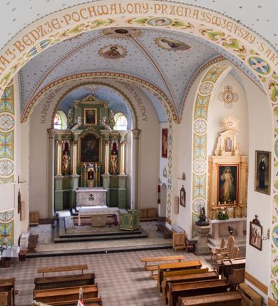 Zdjęcie nr 25: Kościół na planie krzyża łacińskiego, jednonawowy, pięcioprzęsłowy, z transeptem i jednoprzęsłowym prezbiterium zakończonym wielobocznie. Ramiona transeptu jednoprzęsłowe, o wysokości nawy, zamknięte prostymi ścianami. W narożnikach pomiędzy prezbiterium i ramionami transeptu dwa prostokątne aneksy mieszczące zakrystię i skarbiec. Prezbiterium i ramiona transeptu oddzielone od nawy wydatnymi półfilarami przechodzącymi bezpośrednio w gurty sklepienne. Prezbiterium od absydy oddziela arkada tęczowa wsparta na dostawionych do węgarów kolumnach kompozytowych. Sklepienie nad skrzyżowaniem naw krzyżowe, nad pozostałymi przęsłami nawy kolebkowe o przekroju koszowym, nad ramionami transeptu krzyżowe (ostrołukowe), w absydzie ćwierćsferyczne z lunetami. Otwory okienne prostokątne, zamknięte półkoliście, rozglifione do wewnątrz; trzy pary w nawie, dwa na ścianach zamykających ramiona transeptu. Ponad oknami w transepcie dodatkowo po jednym okrągłym oknie wypełnionym rozetą. W prezbiterium okna w kształcie wydłużonego prostokąta o smukłych proporcjach zamkniętego półkoliście, pomiędzy nimi para regularnie rozmieszczonych okien owalnych.  W zachodnim przęśle nawy murowany trójprzęsłowy chór muzyczny wsparty na parze ośmiobocznych filarów z cokołami, a pod ścianami na parze półfilarów. Balustrada o prostej dekoracji płycinowej. Podniebie chóru podzielone belkami na trzy kasetony, pośrodku których kwadratowe płyciny. Fasada kościoła dwukondygnacyjna, trójosiowa przechodząca w czworoboczną wieżę. Ściany rozczłonkowane lizenami, kondygnacje oddzielone profilowanym gzymsem. W części środkowej wejście zamknięte łukiem półkolistym, z dwuspadowym drewnianym daszkiem, wyżej prostokątne okno zamknięte półkoliście. W polach bocznych mniejsze, prostokątne okna zamknięte półkoliście. Wieża o narożach ujętych lizenami, podtrzymującymi gzyms, nakryta ozdobnym zwieńczeniem z czterema wieżyczkami w narożach, pomiędzy którymi trójkątne szczyciki nad oknami i ostrosłupowy hełm zakończony krzyżem. Ściany boczne nawy i transeptu ujęte uskokowymi przyporami. Ściany zamykające ramiona transeptu podzielone na dwie strefy, z których dolna ujęta lizenami, górna wypełniona oknem w ozdobnym obramieniu. Dachy dwuspadowe, kryte dachówką. Na skrzyżowaniu naw wieloboczna, drewniana wieżyczka na sygnaturkę nakryta ostrosłupowym hełmem pokrytym blachą.

