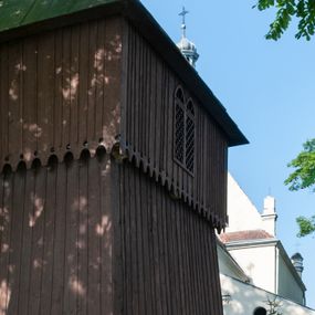 Zdjęcie nr 1: Drewniana dzwonnica została wybudowana 15 m na południowy wschód od kościoła w Wawrzeńczycach, w obrębie ogrodzenia na terenie dawnego cmentarza. Ustawiono ją w konstrukcji słupowej, na kamiennej podmurówce, na planie kwadratu, jako budowlę dwukondygnacyjną z izbicą. Wieża została oszalowana deskami o układzie pionowym. Dolna kondygnacja o ścianach pochyłych. W elewacji północnej dwa prostokątne otwory wejściowe nakryte prostymi daszkami pulpitowymi, mieszczące jednoskrzydłowe, drewniane drzwi. W górnej części każdej elewacji dwudzielne okna zakończone ostrołukowo, wypełnione kratownicą drewnianą. Izbica zakończona od dołu ażurową dekoracją w formie wyciętych w deskach zaokrąglonych zębów oraz okrągłych otworów. Nakryta dachem czterospadowym o konstrukcji krokwiowo-płatwiowej, zabezpieczona blachą ocynkowaną. W środku trzykondygnacyjna, z drewnianymi schodami; mieści trzy, barokowe dzwony.