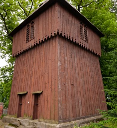 Zdjęcie nr 3: Drewniana dzwonnica została wybudowana 15 m na południowy wschód od kościoła w Wawrzeńczycach, w obrębie ogrodzenia na terenie dawnego cmentarza. Ustawiono ją w konstrukcji słupowej, na kamiennej podmurówce, na planie kwadratu, jako budowlę dwukondygnacyjną z izbicą. Wieża została oszalowana deskami o układzie pionowym. Dolna kondygnacja o ścianach pochyłych. W elewacji północnej dwa prostokątne otwory wejściowe nakryte prostymi daszkami pulpitowymi, mieszczące jednoskrzydłowe, drewniane drzwi. W górnej części każdej elewacji dwudzielne okna zakończone ostrołukowo, wypełnione kratownicą drewnianą. Izbica zakończona od dołu ażurową dekoracją w formie wyciętych w deskach zaokrąglonych zębów oraz okrągłych otworów. Nakryta dachem czterospadowym o konstrukcji krokwiowo-płatwiowej, zabezpieczona blachą ocynkowaną. W środku trzykondygnacyjna, z drewnianymi schodami; mieści trzy, barokowe dzwony.