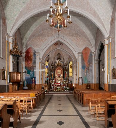 Zdjęcie nr 27: Kościół murowany z cegły, tynkowany, orientowany z odchyleniem ku północy. Zbudowany na planie krzyża łacińskiego, jednonawowy z transeptem, pięcioprzęsłowy, z jednoprzęsłowym prezbiterium zamkniętym trójbocznie. Sklepienie beczkowe spłaszczone (koszowe) na gurtach z lunetami stykającymi się wierzchołkami. W pierwszym od zachodu przęśle nawy chór muzyczny konstrukcji żelbetowej wsparty na dwóch filarach o przekroju ośmioboku i dwóch analogicznych półfilarach. Balustrada artykułowana ostrołukowymi niszami. Ściany boczne rozczłonkowane analogicznymi półfilarami, ustawionymi na tle pilastrów; proste kapitele toskańskie. Ściana tęczowa z arkadą ostrołukową, ujętą po bokach dwoma mniejszymi ostrołukowymi płycinami. Od zachodu prostokątna kruchta z dwoma ostrołukowymi otworami drzwiowymi. Mniejsza prostokątna kruchta od południa, skomunikowana z nawą kamiennym, profilowanym portalem prostokątnym. W prezbiterium od południa prostokątny otwór wejściowy do zakrystii. Okna obustronnie rozglifione, z  parapetami, o kształcie wydłużonego prostokąta, zamkniętego ostrołukowo; po trzy w ścianach bocznych nawy, trzy w prezbiterium. Dwa mniejsze w fasadzie kościoła, po jednym na ścianach bocznych kruchty zachodniej. W zamknięciu ścian transeptu, większe ostrołukowe okna z podziałami kamiennymi dzielącymi okno na dwie, mniejsze, przedzielone półfilarem, zamknięte ostrołukowo części. Kościół nakryty dachem dwuspadowym z blachy. Fasada bezwieżowa z trójkątnym szczytem. Na zewnętrznej, wschodniej ścianie prezbiterium niewielka kwadratowa płycina z inskrypcją: „KAMIEŃ / WĘGIELNY / POŚWIĘCONY / DN(IA) 21 VI 1936”.