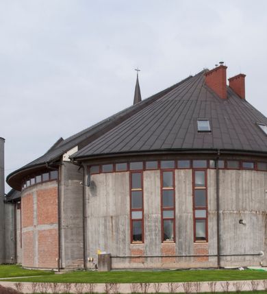 Zdjęcie nr 3: Kościół usytuowany na dużej działce o nieregularnym kształcie.  Świątynia otoczona trawnikiem, w którym wydzielono niewielkie klomby kwiatowe i ciągi komunikacyjne. Kościół orientowany, jednonawowy, jednoprzestrzenny z prezbiterium zamkniętym od zewnątrz szeroką absydą. Ściany boczne nawy od strony wschodniej założone na planie odcinka koła. Wnętrze jednoprzestrzenne, z widoczną konstrukcją otwartej więźby dachowej. Widoczne, drewniane belki wspierające się na 12 kolumnach, ustawionych w dwa rzędy, w układzie powtarzających rytm ścian. Prezbiterium na trójstopniowym wzniesieniu, oddzielone od nawy ścianą tęczową, zamknięte prostą ścianą. Od strony zachodniej chór muzyczny konstrukcji żelbetowej o pełnym, prostym parapecie, wsparty na dwóch okrągłych filarach. Po cztery pionowe okna łączące się w górnych partiach z świetlikiem obiegającym całą nawę. Mury zakończone uproszczonym gzymsem. W północno-zachodnim narożu niewielka kaplica Jana Pawła II, założona na planie koła. Fasada kościoła z trójkątną ścianą szczytową wyłożoną okładziną kamienną z umieszczonym w osi dużym okrągłym oknem, ujęta dwoma betonowymi wieżami na planie koła. W górnej części wieże z prześwitami na dzwony, nakryte strzelistymi, ostrosłupowymi dachami. W przyziemiu portyk wejściowy w formie czterech kolumn dźwigających masywny gzyms z napisem: „KOŚCIÓŁ MATKI BOSKIEJ RÓŻAŃCOWEJ”, powyżej rzeźba Madonny z Dzieciątkiem. 

