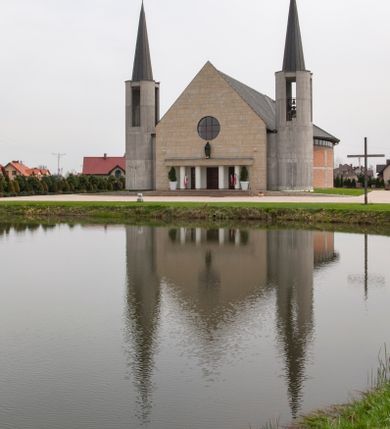 Zdjęcie nr 4: Kościół usytuowany na dużej działce o nieregularnym kształcie.  Świątynia otoczona trawnikiem, w którym wydzielono niewielkie klomby kwiatowe i ciągi komunikacyjne. Kościół orientowany, jednonawowy, jednoprzestrzenny z prezbiterium zamkniętym od zewnątrz szeroką absydą. Ściany boczne nawy od strony wschodniej założone na planie odcinka koła. Wnętrze jednoprzestrzenne, z widoczną konstrukcją otwartej więźby dachowej. Widoczne, drewniane belki wspierające się na 12 kolumnach, ustawionych w dwa rzędy, w układzie powtarzających rytm ścian. Prezbiterium na trójstopniowym wzniesieniu, oddzielone od nawy ścianą tęczową, zamknięte prostą ścianą. Od strony zachodniej chór muzyczny konstrukcji żelbetowej o pełnym, prostym parapecie, wsparty na dwóch okrągłych filarach. Po cztery pionowe okna łączące się w górnych partiach z świetlikiem obiegającym całą nawę. Mury zakończone uproszczonym gzymsem. W północno-zachodnim narożu niewielka kaplica Jana Pawła II, założona na planie koła. Fasada kościoła z trójkątną ścianą szczytową wyłożoną okładziną kamienną z umieszczonym w osi dużym okrągłym oknem, ujęta dwoma betonowymi wieżami na planie koła. W górnej części wieże z prześwitami na dzwony, nakryte strzelistymi, ostrosłupowymi dachami. W przyziemiu portyk wejściowy w formie czterech kolumn dźwigających masywny gzyms z napisem: „KOŚCIÓŁ MATKI BOSKIEJ RÓŻAŃCOWEJ”, powyżej rzeźba Madonny z Dzieciątkiem. 

