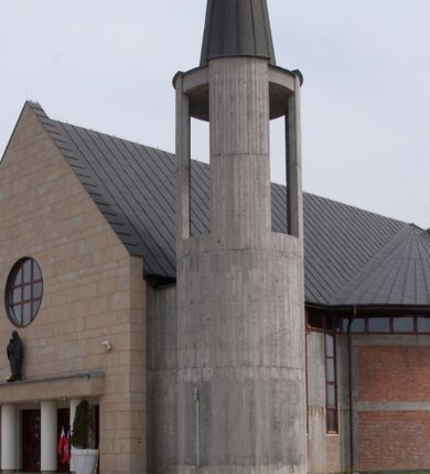 Zdjęcie nr 1: Kościół usytuowany na dużej działce o nieregularnym kształcie.  Świątynia otoczona trawnikiem, w którym wydzielono niewielkie klomby kwiatowe i ciągi komunikacyjne. Kościół orientowany, jednonawowy, jednoprzestrzenny z prezbiterium zamkniętym od zewnątrz szeroką absydą. Ściany boczne nawy od strony wschodniej założone na planie odcinka koła. Wnętrze jednoprzestrzenne, z widoczną konstrukcją otwartej więźby dachowej. Widoczne, drewniane belki wspierające się na 12 kolumnach, ustawionych w dwa rzędy, w układzie powtarzających rytm ścian. Prezbiterium na trójstopniowym wzniesieniu, oddzielone od nawy ścianą tęczową, zamknięte prostą ścianą. Od strony zachodniej chór muzyczny konstrukcji żelbetowej o pełnym, prostym parapecie, wsparty na dwóch okrągłych filarach. Po cztery pionowe okna łączące się w górnych partiach z świetlikiem obiegającym całą nawę. Mury zakończone uproszczonym gzymsem. W północno-zachodnim narożu niewielka kaplica Jana Pawła II, założona na planie koła. Fasada kościoła z trójkątną ścianą szczytową wyłożoną okładziną kamienną z umieszczonym w osi dużym okrągłym oknem, ujęta dwoma betonowymi wieżami na planie koła. W górnej części wieże z prześwitami na dzwony, nakryte strzelistymi, ostrosłupowymi dachami. W przyziemiu portyk wejściowy w formie czterech kolumn dźwigających masywny gzyms z napisem: „KOŚCIÓŁ MATKI BOSKIEJ RÓŻAŃCOWEJ”, powyżej rzeźba Madonny z Dzieciątkiem. 

