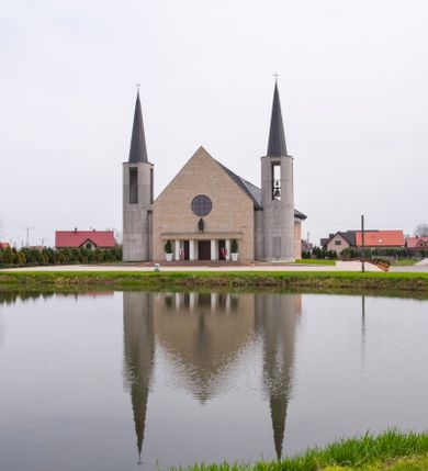 Zdjęcie nr 8: Kościół usytuowany na dużej działce o nieregularnym kształcie.  Świątynia otoczona trawnikiem, w którym wydzielono niewielkie klomby kwiatowe i ciągi komunikacyjne. Kościół orientowany, jednonawowy, jednoprzestrzenny z prezbiterium zamkniętym od zewnątrz szeroką absydą. Ściany boczne nawy od strony wschodniej założone na planie odcinka koła. Wnętrze jednoprzestrzenne, z widoczną konstrukcją otwartej więźby dachowej. Widoczne, drewniane belki wspierające się na 12 kolumnach, ustawionych w dwa rzędy, w układzie powtarzających rytm ścian. Prezbiterium na trójstopniowym wzniesieniu, oddzielone od nawy ścianą tęczową, zamknięte prostą ścianą. Od strony zachodniej chór muzyczny konstrukcji żelbetowej o pełnym, prostym parapecie, wsparty na dwóch okrągłych filarach. Po cztery pionowe okna łączące się w górnych partiach z świetlikiem obiegającym całą nawę. Mury zakończone uproszczonym gzymsem. W północno-zachodnim narożu niewielka kaplica Jana Pawła II, założona na planie koła. Fasada kościoła z trójkątną ścianą szczytową wyłożoną okładziną kamienną z umieszczonym w osi dużym okrągłym oknem, ujęta dwoma betonowymi wieżami na planie koła. W górnej części wieże z prześwitami na dzwony, nakryte strzelistymi, ostrosłupowymi dachami. W przyziemiu portyk wejściowy w formie czterech kolumn dźwigających masywny gzyms z napisem: „KOŚCIÓŁ MATKI BOSKIEJ RÓŻAŃCOWEJ”, powyżej rzeźba Madonny z Dzieciątkiem. 

