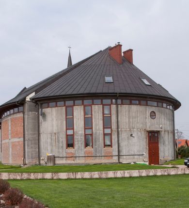 Zdjęcie nr 5: Kościół usytuowany na dużej działce o nieregularnym kształcie.  Świątynia otoczona trawnikiem, w którym wydzielono niewielkie klomby kwiatowe i ciągi komunikacyjne. Kościół orientowany, jednonawowy, jednoprzestrzenny z prezbiterium zamkniętym od zewnątrz szeroką absydą. Ściany boczne nawy od strony wschodniej założone na planie odcinka koła. Wnętrze jednoprzestrzenne, z widoczną konstrukcją otwartej więźby dachowej. Widoczne, drewniane belki wspierające się na 12 kolumnach, ustawionych w dwa rzędy, w układzie powtarzających rytm ścian. Prezbiterium na trójstopniowym wzniesieniu, oddzielone od nawy ścianą tęczową, zamknięte prostą ścianą. Od strony zachodniej chór muzyczny konstrukcji żelbetowej o pełnym, prostym parapecie, wsparty na dwóch okrągłych filarach. Po cztery pionowe okna łączące się w górnych partiach z świetlikiem obiegającym całą nawę. Mury zakończone uproszczonym gzymsem. W północno-zachodnim narożu niewielka kaplica Jana Pawła II, założona na planie koła. Fasada kościoła z trójkątną ścianą szczytową wyłożoną okładziną kamienną z umieszczonym w osi dużym okrągłym oknem, ujęta dwoma betonowymi wieżami na planie koła. W górnej części wieże z prześwitami na dzwony, nakryte strzelistymi, ostrosłupowymi dachami. W przyziemiu portyk wejściowy w formie czterech kolumn dźwigających masywny gzyms z napisem: „KOŚCIÓŁ MATKI BOSKIEJ RÓŻAŃCOWEJ”, powyżej rzeźba Madonny z Dzieciątkiem. 

