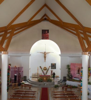 Zdjęcie nr 12: Kościół usytuowany na dużej działce o nieregularnym kształcie.  Świątynia otoczona trawnikiem, w którym wydzielono niewielkie klomby kwiatowe i ciągi komunikacyjne. Kościół orientowany, jednonawowy, jednoprzestrzenny z prezbiterium zamkniętym od zewnątrz szeroką absydą. Ściany boczne nawy od strony wschodniej założone na planie odcinka koła. Wnętrze jednoprzestrzenne, z widoczną konstrukcją otwartej więźby dachowej. Widoczne, drewniane belki wspierające się na 12 kolumnach, ustawionych w dwa rzędy, w układzie powtarzających rytm ścian. Prezbiterium na trójstopniowym wzniesieniu, oddzielone od nawy ścianą tęczową, zamknięte prostą ścianą. Od strony zachodniej chór muzyczny konstrukcji żelbetowej o pełnym, prostym parapecie, wsparty na dwóch okrągłych filarach. Po cztery pionowe okna łączące się w górnych partiach z świetlikiem obiegającym całą nawę. Mury zakończone uproszczonym gzymsem. W północno-zachodnim narożu niewielka kaplica Jana Pawła II, założona na planie koła. Fasada kościoła z trójkątną ścianą szczytową wyłożoną okładziną kamienną z umieszczonym w osi dużym okrągłym oknem, ujęta dwoma betonowymi wieżami na planie koła. W górnej części wieże z prześwitami na dzwony, nakryte strzelistymi, ostrosłupowymi dachami. W przyziemiu portyk wejściowy w formie czterech kolumn dźwigających masywny gzyms z napisem: „KOŚCIÓŁ MATKI BOSKIEJ RÓŻAŃCOWEJ”, powyżej rzeźba Madonny z Dzieciątkiem. 

