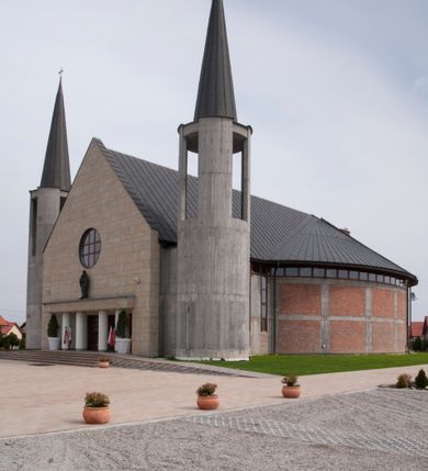 Zdjęcie nr 7: Kościół usytuowany na dużej działce o nieregularnym kształcie.  Świątynia otoczona trawnikiem, w którym wydzielono niewielkie klomby kwiatowe i ciągi komunikacyjne. Kościół orientowany, jednonawowy, jednoprzestrzenny z prezbiterium zamkniętym od zewnątrz szeroką absydą. Ściany boczne nawy od strony wschodniej założone na planie odcinka koła. Wnętrze jednoprzestrzenne, z widoczną konstrukcją otwartej więźby dachowej. Widoczne, drewniane belki wspierające się na 12 kolumnach, ustawionych w dwa rzędy, w układzie powtarzających rytm ścian. Prezbiterium na trójstopniowym wzniesieniu, oddzielone od nawy ścianą tęczową, zamknięte prostą ścianą. Od strony zachodniej chór muzyczny konstrukcji żelbetowej o pełnym, prostym parapecie, wsparty na dwóch okrągłych filarach. Po cztery pionowe okna łączące się w górnych partiach z świetlikiem obiegającym całą nawę. Mury zakończone uproszczonym gzymsem. W północno-zachodnim narożu niewielka kaplica Jana Pawła II, założona na planie koła. Fasada kościoła z trójkątną ścianą szczytową wyłożoną okładziną kamienną z umieszczonym w osi dużym okrągłym oknem, ujęta dwoma betonowymi wieżami na planie koła. W górnej części wieże z prześwitami na dzwony, nakryte strzelistymi, ostrosłupowymi dachami. W przyziemiu portyk wejściowy w formie czterech kolumn dźwigających masywny gzyms z napisem: „KOŚCIÓŁ MATKI BOSKIEJ RÓŻAŃCOWEJ”, powyżej rzeźba Madonny z Dzieciątkiem. 

