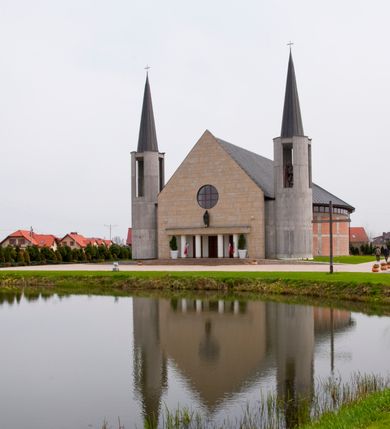 Zdjęcie nr 10: Kościół usytuowany na dużej działce o nieregularnym kształcie.  Świątynia otoczona trawnikiem, w którym wydzielono niewielkie klomby kwiatowe i ciągi komunikacyjne. Kościół orientowany, jednonawowy, jednoprzestrzenny z prezbiterium zamkniętym od zewnątrz szeroką absydą. Ściany boczne nawy od strony wschodniej założone na planie odcinka koła. Wnętrze jednoprzestrzenne, z widoczną konstrukcją otwartej więźby dachowej. Widoczne, drewniane belki wspierające się na 12 kolumnach, ustawionych w dwa rzędy, w układzie powtarzających rytm ścian. Prezbiterium na trójstopniowym wzniesieniu, oddzielone od nawy ścianą tęczową, zamknięte prostą ścianą. Od strony zachodniej chór muzyczny konstrukcji żelbetowej o pełnym, prostym parapecie, wsparty na dwóch okrągłych filarach. Po cztery pionowe okna łączące się w górnych partiach z świetlikiem obiegającym całą nawę. Mury zakończone uproszczonym gzymsem. W północno-zachodnim narożu niewielka kaplica Jana Pawła II, założona na planie koła. Fasada kościoła z trójkątną ścianą szczytową wyłożoną okładziną kamienną z umieszczonym w osi dużym okrągłym oknem, ujęta dwoma betonowymi wieżami na planie koła. W górnej części wieże z prześwitami na dzwony, nakryte strzelistymi, ostrosłupowymi dachami. W przyziemiu portyk wejściowy w formie czterech kolumn dźwigających masywny gzyms z napisem: „KOŚCIÓŁ MATKI BOSKIEJ RÓŻAŃCOWEJ”, powyżej rzeźba Madonny z Dzieciątkiem. 

