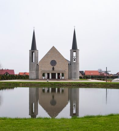 Zdjęcie nr 9: Kościół usytuowany na dużej działce o nieregularnym kształcie.  Świątynia otoczona trawnikiem, w którym wydzielono niewielkie klomby kwiatowe i ciągi komunikacyjne. Kościół orientowany, jednonawowy, jednoprzestrzenny z prezbiterium zamkniętym od zewnątrz szeroką absydą. Ściany boczne nawy od strony wschodniej założone na planie odcinka koła. Wnętrze jednoprzestrzenne, z widoczną konstrukcją otwartej więźby dachowej. Widoczne, drewniane belki wspierające się na 12 kolumnach, ustawionych w dwa rzędy, w układzie powtarzających rytm ścian. Prezbiterium na trójstopniowym wzniesieniu, oddzielone od nawy ścianą tęczową, zamknięte prostą ścianą. Od strony zachodniej chór muzyczny konstrukcji żelbetowej o pełnym, prostym parapecie, wsparty na dwóch okrągłych filarach. Po cztery pionowe okna łączące się w górnych partiach z świetlikiem obiegającym całą nawę. Mury zakończone uproszczonym gzymsem. W północno-zachodnim narożu niewielka kaplica Jana Pawła II, założona na planie koła. Fasada kościoła z trójkątną ścianą szczytową wyłożoną okładziną kamienną z umieszczonym w osi dużym okrągłym oknem, ujęta dwoma betonowymi wieżami na planie koła. W górnej części wieże z prześwitami na dzwony, nakryte strzelistymi, ostrosłupowymi dachami. W przyziemiu portyk wejściowy w formie czterech kolumn dźwigających masywny gzyms z napisem: „KOŚCIÓŁ MATKI BOSKIEJ RÓŻAŃCOWEJ”, powyżej rzeźba Madonny z Dzieciątkiem. 

