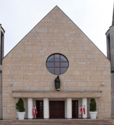 Zdjęcie nr 6: Kościół usytuowany na dużej działce o nieregularnym kształcie.  Świątynia otoczona trawnikiem, w którym wydzielono niewielkie klomby kwiatowe i ciągi komunikacyjne. Kościół orientowany, jednonawowy, jednoprzestrzenny z prezbiterium zamkniętym od zewnątrz szeroką absydą. Ściany boczne nawy od strony wschodniej założone na planie odcinka koła. Wnętrze jednoprzestrzenne, z widoczną konstrukcją otwartej więźby dachowej. Widoczne, drewniane belki wspierające się na 12 kolumnach, ustawionych w dwa rzędy, w układzie powtarzających rytm ścian. Prezbiterium na trójstopniowym wzniesieniu, oddzielone od nawy ścianą tęczową, zamknięte prostą ścianą. Od strony zachodniej chór muzyczny konstrukcji żelbetowej o pełnym, prostym parapecie, wsparty na dwóch okrągłych filarach. Po cztery pionowe okna łączące się w górnych partiach z świetlikiem obiegającym całą nawę. Mury zakończone uproszczonym gzymsem. W północno-zachodnim narożu niewielka kaplica Jana Pawła II, założona na planie koła. Fasada kościoła z trójkątną ścianą szczytową wyłożoną okładziną kamienną z umieszczonym w osi dużym okrągłym oknem, ujęta dwoma betonowymi wieżami na planie koła. W górnej części wieże z prześwitami na dzwony, nakryte strzelistymi, ostrosłupowymi dachami. W przyziemiu portyk wejściowy w formie czterech kolumn dźwigających masywny gzyms z napisem: „KOŚCIÓŁ MATKI BOSKIEJ RÓŻAŃCOWEJ”, powyżej rzeźba Madonny z Dzieciątkiem. 

