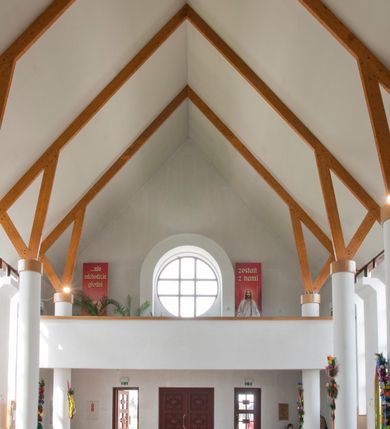 Zdjęcie nr 25: Kościół usytuowany na dużej działce o nieregularnym kształcie.  Świątynia otoczona trawnikiem, w którym wydzielono niewielkie klomby kwiatowe i ciągi komunikacyjne. Kościół orientowany, jednonawowy, jednoprzestrzenny z prezbiterium zamkniętym od zewnątrz szeroką absydą. Ściany boczne nawy od strony wschodniej założone na planie odcinka koła. Wnętrze jednoprzestrzenne, z widoczną konstrukcją otwartej więźby dachowej. Widoczne, drewniane belki wspierające się na 12 kolumnach, ustawionych w dwa rzędy, w układzie powtarzających rytm ścian. Prezbiterium na trójstopniowym wzniesieniu, oddzielone od nawy ścianą tęczową, zamknięte prostą ścianą. Od strony zachodniej chór muzyczny konstrukcji żelbetowej o pełnym, prostym parapecie, wsparty na dwóch okrągłych filarach. Po cztery pionowe okna łączące się w górnych partiach z świetlikiem obiegającym całą nawę. Mury zakończone uproszczonym gzymsem. W północno-zachodnim narożu niewielka kaplica Jana Pawła II, założona na planie koła. Fasada kościoła z trójkątną ścianą szczytową wyłożoną okładziną kamienną z umieszczonym w osi dużym okrągłym oknem, ujęta dwoma betonowymi wieżami na planie koła. W górnej części wieże z prześwitami na dzwony, nakryte strzelistymi, ostrosłupowymi dachami. W przyziemiu portyk wejściowy w formie czterech kolumn dźwigających masywny gzyms z napisem: „KOŚCIÓŁ MATKI BOSKIEJ RÓŻAŃCOWEJ”, powyżej rzeźba Madonny z Dzieciątkiem. 

