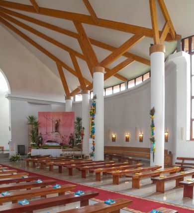 Zdjęcie nr 15: Kościół usytuowany na dużej działce o nieregularnym kształcie.  Świątynia otoczona trawnikiem, w którym wydzielono niewielkie klomby kwiatowe i ciągi komunikacyjne. Kościół orientowany, jednonawowy, jednoprzestrzenny z prezbiterium zamkniętym od zewnątrz szeroką absydą. Ściany boczne nawy od strony wschodniej założone na planie odcinka koła. Wnętrze jednoprzestrzenne, z widoczną konstrukcją otwartej więźby dachowej. Widoczne, drewniane belki wspierające się na 12 kolumnach, ustawionych w dwa rzędy, w układzie powtarzających rytm ścian. Prezbiterium na trójstopniowym wzniesieniu, oddzielone od nawy ścianą tęczową, zamknięte prostą ścianą. Od strony zachodniej chór muzyczny konstrukcji żelbetowej o pełnym, prostym parapecie, wsparty na dwóch okrągłych filarach. Po cztery pionowe okna łączące się w górnych partiach z świetlikiem obiegającym całą nawę. Mury zakończone uproszczonym gzymsem. W północno-zachodnim narożu niewielka kaplica Jana Pawła II, założona na planie koła. Fasada kościoła z trójkątną ścianą szczytową wyłożoną okładziną kamienną z umieszczonym w osi dużym okrągłym oknem, ujęta dwoma betonowymi wieżami na planie koła. W górnej części wieże z prześwitami na dzwony, nakryte strzelistymi, ostrosłupowymi dachami. W przyziemiu portyk wejściowy w formie czterech kolumn dźwigających masywny gzyms z napisem: „KOŚCIÓŁ MATKI BOSKIEJ RÓŻAŃCOWEJ”, powyżej rzeźba Madonny z Dzieciątkiem. 

