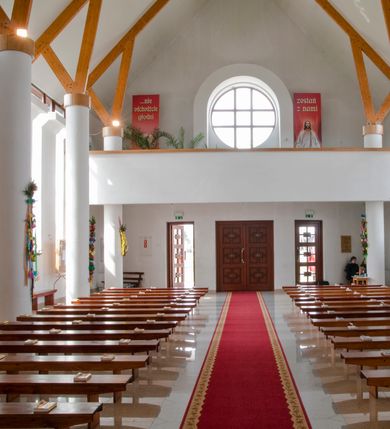 Zdjęcie nr 14: Kościół usytuowany na dużej działce o nieregularnym kształcie.  Świątynia otoczona trawnikiem, w którym wydzielono niewielkie klomby kwiatowe i ciągi komunikacyjne. Kościół orientowany, jednonawowy, jednoprzestrzenny z prezbiterium zamkniętym od zewnątrz szeroką absydą. Ściany boczne nawy od strony wschodniej założone na planie odcinka koła. Wnętrze jednoprzestrzenne, z widoczną konstrukcją otwartej więźby dachowej. Widoczne, drewniane belki wspierające się na 12 kolumnach, ustawionych w dwa rzędy, w układzie powtarzających rytm ścian. Prezbiterium na trójstopniowym wzniesieniu, oddzielone od nawy ścianą tęczową, zamknięte prostą ścianą. Od strony zachodniej chór muzyczny konstrukcji żelbetowej o pełnym, prostym parapecie, wsparty na dwóch okrągłych filarach. Po cztery pionowe okna łączące się w górnych partiach z świetlikiem obiegającym całą nawę. Mury zakończone uproszczonym gzymsem. W północno-zachodnim narożu niewielka kaplica Jana Pawła II, założona na planie koła. Fasada kościoła z trójkątną ścianą szczytową wyłożoną okładziną kamienną z umieszczonym w osi dużym okrągłym oknem, ujęta dwoma betonowymi wieżami na planie koła. W górnej części wieże z prześwitami na dzwony, nakryte strzelistymi, ostrosłupowymi dachami. W przyziemiu portyk wejściowy w formie czterech kolumn dźwigających masywny gzyms z napisem: „KOŚCIÓŁ MATKI BOSKIEJ RÓŻAŃCOWEJ”, powyżej rzeźba Madonny z Dzieciątkiem. 

