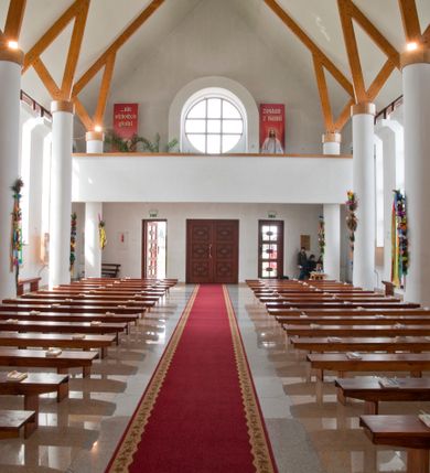 Zdjęcie nr 17: Kościół usytuowany na dużej działce o nieregularnym kształcie.  Świątynia otoczona trawnikiem, w którym wydzielono niewielkie klomby kwiatowe i ciągi komunikacyjne. Kościół orientowany, jednonawowy, jednoprzestrzenny z prezbiterium zamkniętym od zewnątrz szeroką absydą. Ściany boczne nawy od strony wschodniej założone na planie odcinka koła. Wnętrze jednoprzestrzenne, z widoczną konstrukcją otwartej więźby dachowej. Widoczne, drewniane belki wspierające się na 12 kolumnach, ustawionych w dwa rzędy, w układzie powtarzających rytm ścian. Prezbiterium na trójstopniowym wzniesieniu, oddzielone od nawy ścianą tęczową, zamknięte prostą ścianą. Od strony zachodniej chór muzyczny konstrukcji żelbetowej o pełnym, prostym parapecie, wsparty na dwóch okrągłych filarach. Po cztery pionowe okna łączące się w górnych partiach z świetlikiem obiegającym całą nawę. Mury zakończone uproszczonym gzymsem. W północno-zachodnim narożu niewielka kaplica Jana Pawła II, założona na planie koła. Fasada kościoła z trójkątną ścianą szczytową wyłożoną okładziną kamienną z umieszczonym w osi dużym okrągłym oknem, ujęta dwoma betonowymi wieżami na planie koła. W górnej części wieże z prześwitami na dzwony, nakryte strzelistymi, ostrosłupowymi dachami. W przyziemiu portyk wejściowy w formie czterech kolumn dźwigających masywny gzyms z napisem: „KOŚCIÓŁ MATKI BOSKIEJ RÓŻAŃCOWEJ”, powyżej rzeźba Madonny z Dzieciątkiem. 

