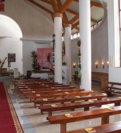 Zdjęcie nr 18: Kościół usytuowany na dużej działce o nieregularnym kształcie.  Świątynia otoczona trawnikiem, w którym wydzielono niewielkie klomby kwiatowe i ciągi komunikacyjne. Kościół orientowany, jednonawowy, jednoprzestrzenny z prezbiterium zamkniętym od zewnątrz szeroką absydą. Ściany boczne nawy od strony wschodniej założone na planie odcinka koła. Wnętrze jednoprzestrzenne, z widoczną konstrukcją otwartej więźby dachowej. Widoczne, drewniane belki wspierające się na 12 kolumnach, ustawionych w dwa rzędy, w układzie powtarzających rytm ścian. Prezbiterium na trójstopniowym wzniesieniu, oddzielone od nawy ścianą tęczową, zamknięte prostą ścianą. Od strony zachodniej chór muzyczny konstrukcji żelbetowej o pełnym, prostym parapecie, wsparty na dwóch okrągłych filarach. Po cztery pionowe okna łączące się w górnych partiach z świetlikiem obiegającym całą nawę. Mury zakończone uproszczonym gzymsem. W północno-zachodnim narożu niewielka kaplica Jana Pawła II, założona na planie koła. Fasada kościoła z trójkątną ścianą szczytową wyłożoną okładziną kamienną z umieszczonym w osi dużym okrągłym oknem, ujęta dwoma betonowymi wieżami na planie koła. W górnej części wieże z prześwitami na dzwony, nakryte strzelistymi, ostrosłupowymi dachami. W przyziemiu portyk wejściowy w formie czterech kolumn dźwigających masywny gzyms z napisem: „KOŚCIÓŁ MATKI BOSKIEJ RÓŻAŃCOWEJ”, powyżej rzeźba Madonny z Dzieciątkiem. 

