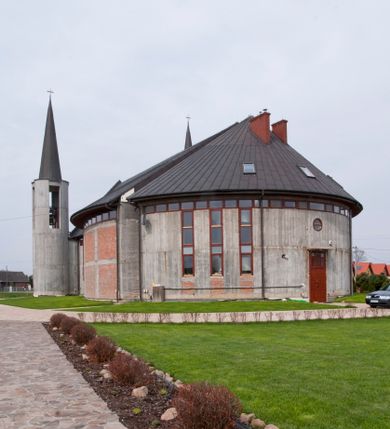 Zdjęcie nr 11: Kościół usytuowany na dużej działce o nieregularnym kształcie.  Świątynia otoczona trawnikiem, w którym wydzielono niewielkie klomby kwiatowe i ciągi komunikacyjne. Kościół orientowany, jednonawowy, jednoprzestrzenny z prezbiterium zamkniętym od zewnątrz szeroką absydą. Ściany boczne nawy od strony wschodniej założone na planie odcinka koła. Wnętrze jednoprzestrzenne, z widoczną konstrukcją otwartej więźby dachowej. Widoczne, drewniane belki wspierające się na 12 kolumnach, ustawionych w dwa rzędy, w układzie powtarzających rytm ścian. Prezbiterium na trójstopniowym wzniesieniu, oddzielone od nawy ścianą tęczową, zamknięte prostą ścianą. Od strony zachodniej chór muzyczny konstrukcji żelbetowej o pełnym, prostym parapecie, wsparty na dwóch okrągłych filarach. Po cztery pionowe okna łączące się w górnych partiach z świetlikiem obiegającym całą nawę. Mury zakończone uproszczonym gzymsem. W północno-zachodnim narożu niewielka kaplica Jana Pawła II, założona na planie koła. Fasada kościoła z trójkątną ścianą szczytową wyłożoną okładziną kamienną z umieszczonym w osi dużym okrągłym oknem, ujęta dwoma betonowymi wieżami na planie koła. W górnej części wieże z prześwitami na dzwony, nakryte strzelistymi, ostrosłupowymi dachami. W przyziemiu portyk wejściowy w formie czterech kolumn dźwigających masywny gzyms z napisem: „KOŚCIÓŁ MATKI BOSKIEJ RÓŻAŃCOWEJ”, powyżej rzeźba Madonny z Dzieciątkiem. 

