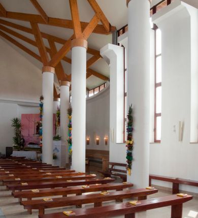 Zdjęcie nr 22: Kościół usytuowany na dużej działce o nieregularnym kształcie.  Świątynia otoczona trawnikiem, w którym wydzielono niewielkie klomby kwiatowe i ciągi komunikacyjne. Kościół orientowany, jednonawowy, jednoprzestrzenny z prezbiterium zamkniętym od zewnątrz szeroką absydą. Ściany boczne nawy od strony wschodniej założone na planie odcinka koła. Wnętrze jednoprzestrzenne, z widoczną konstrukcją otwartej więźby dachowej. Widoczne, drewniane belki wspierające się na 12 kolumnach, ustawionych w dwa rzędy, w układzie powtarzających rytm ścian. Prezbiterium na trójstopniowym wzniesieniu, oddzielone od nawy ścianą tęczową, zamknięte prostą ścianą. Od strony zachodniej chór muzyczny konstrukcji żelbetowej o pełnym, prostym parapecie, wsparty na dwóch okrągłych filarach. Po cztery pionowe okna łączące się w górnych partiach z świetlikiem obiegającym całą nawę. Mury zakończone uproszczonym gzymsem. W północno-zachodnim narożu niewielka kaplica Jana Pawła II, założona na planie koła. Fasada kościoła z trójkątną ścianą szczytową wyłożoną okładziną kamienną z umieszczonym w osi dużym okrągłym oknem, ujęta dwoma betonowymi wieżami na planie koła. W górnej części wieże z prześwitami na dzwony, nakryte strzelistymi, ostrosłupowymi dachami. W przyziemiu portyk wejściowy w formie czterech kolumn dźwigających masywny gzyms z napisem: „KOŚCIÓŁ MATKI BOSKIEJ RÓŻAŃCOWEJ”, powyżej rzeźba Madonny z Dzieciątkiem. 

