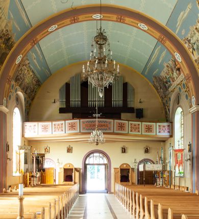 Zdjęcie nr 10: Kościół murowany, na kamiennym podmurowaniu, jednonawowy, na planie krzyża. Nawa na rzucie wydłużonego prostokąta, z węższym, jednoprzęsłowym prezbiterium zamkniętym trójbocznie, flankowanym dwoma kwadratowymi aneksami – zakrystią i kaplicą. Od południowego-zachodu dostawiona wieża na planie kwadratu z uskokowym zwężeniem ostatniej kondygnacji, z kruchtą w przyziemiu, ujęta dwiema wieżyczkami na planie ściętego koła, w południowej kręcone schody, w północnej schowek oraz dwoma otwartymi na zewnątrz arkadą aneksami na planie kwadratu z wejściami bocznymi do świątyni. Wnętrze nakryte sklepieniem kolebkowym z dwoma gurtami opadającymi na cztery półfilary. Otwór tęczowy w formie leżącego prostokąta zamkniętego łukiem półkolistym. W południowo-zachodniej części nawy zawieszony parapet chóru muzycznego o wybrzuszonej części środkowej balustrady. Posadzka betonowa, pokryta płytami kamiennymi. Fasada wieżowa, bez podziału na kondygnacje, w narożach ujęta kamiennymi szkarpami. Na osi portal wejściowy w formie kwadratu zamkniętego łukiem półkolistym, z dwuskrzydłowymi drzwiami, nakryty daszkiem okapowym. Nad nim okno witrażowe w formie koła. Wieża przepruta z trzech stron oknami w formie wydłużonego prostokąta zamkniętego półkoliście oraz w najwyższej części z czterech stron potrójnymi, arkadkowymi oknami. Wieżyczki przeprute niewielkimi, okrągłymi okienkami. Elewacje północna i południowa, ujęte we frontowych narożach kamiennymi szkarpami, przeprute trzema oknami witrażowymi w formie wydłużonego prostokąta zamkniętego półkoliście. Kaplica i zakrystia z jednym oknem o kształcie kwadratu zamkniętego łukiem półkolistym. Elewacja północno-wschodnia z dwoma oknami. Wieża nakryta dachem wieżowym kopulastym z latarnią zakończoną krzyżem, wieżyczki daszkami stożkowymi, a aneksy pulpitowymi. Dach nad nawą dwuspadowy z sześcioboczną wieżyczką na sygnaturkę nakrytą daszkiem wieżowym, prezbiterium niższe, przykryte dachem wielospadowym, kaplica i zakrystia dwuspadowym. 