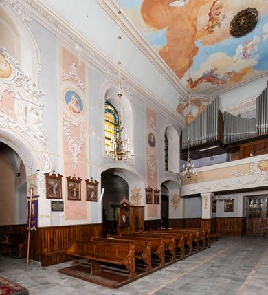 Zdjęcie nr 19: Kościół orientowany, murowany, trójnawowy, z prezbiterium zamkniętym trójbocznie, zakrystią od północy i kruchtą od południa. Do naw przylegają kaplice: większa od północy i mniejsza od południa. Dwie kolejne kaplice przylegają do boków wieży. Ściany wnętrza gładkie, pokryte polichromią imitującą podziały i obramienia, dekorację ornamentalną i sceny iluzjonistyczne na stropach, utrzymane w chłodnej biało-niebiesko-różowej tonacji z dodatkiem żółtego. Na stropie prezbiterium scena Zwiastowania, na stropie nawy Zesłania Ducha Świętego. Od zachodu chór muzyczny wsparty na dwóch filarach z balustradą pokrytą polichromią. Prezbiterium i nawa główna nakryte stropami, nad prezbiterium o przekroju spłaszczonej kolebki, nad nawą płaski, z szerokimi fasetami, zakrystia sklepiona kolebkowo, kruchta i kaplice wschodnie krzyżowo. Nawę, prezbiterium i część kaplic doświetlają okna w kształcie stojącego prostokąta zamkniętego łukiem pełnym, pozostałe zamknięte łukiem odcinkowym lub kwadratowe. Główne wejście i dwa boczne od zachodu, kolejne do przedsionka od północy i do kruchty od południa. Fasada z wieżą na osi, poprzedzona portykiem wspartym na filarach, z trójkątnym szczytem zwieńczonym krzyżem łacińskim. Fasada jednokondygnacyjna, przechodząca w masyw wieży. Kondygnację przepruwają trzy okna i jest zamknięta gzymsem wyniesionym na środku w łuk. Wieńczący ją trójkątny szczyt wtapia się w kondygnację wieży o gładkich elewacjach przeprutych jedynie niewielkimi oknami i tarczami zegarów. Ostatnia kondygnacja jest ujęta gzymsami i dekorowana fryzem arkadowym, którego arkady wykonano z nietynkowanej cegły. Zwieńczenie rozbudowane składa się z iglicy i ustawionych wokół niej, w narożach małych, dwukondygnacyjnych wieżyczek zamkniętych dachami stożkowymi. Wieżyczki połączono podwójnymi arkadami, których łuki wykonano z nietynkowanej cegły, tak samo jak górne kondygnacje wieżyczek. Elewacje kościoła gładkie, na niskim, kamiennym cokole, od wschodu opięte szkarpami. Prezbiterium i nawę przekrywają osobne dachy dwuspadowe, na kalenicy nawy wieżyczka na sygnaturkę, cztery kaplice i portyk nakrywają dachy trójpołaciowe, zakrystię i nawy boczne pulpitowe. 
Jedyne zachowane elementy wyposażenia sprzed pożaru w 1815 roku to fragment osiemnastowiecznej polichromii przedstawiającej dwóch aniołów z narzędziami męki i przedmioty nawiązujące do tego wydarzenia oraz gotycki portal do zakrystii wraz z żelaznymi, kutymi drzwiami. 

