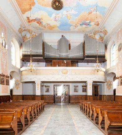 Zdjęcie nr 22: Kościół orientowany, murowany, trójnawowy, z prezbiterium zamkniętym trójbocznie, zakrystią od północy i kruchtą od południa. Do naw przylegają kaplice: większa od północy i mniejsza od południa. Dwie kolejne kaplice przylegają do boków wieży. Ściany wnętrza gładkie, pokryte polichromią imitującą podziały i obramienia, dekorację ornamentalną i sceny iluzjonistyczne na stropach, utrzymane w chłodnej biało-niebiesko-różowej tonacji z dodatkiem żółtego. Na stropie prezbiterium scena Zwiastowania, na stropie nawy Zesłania Ducha Świętego. Od zachodu chór muzyczny wsparty na dwóch filarach z balustradą pokrytą polichromią. Prezbiterium i nawa główna nakryte stropami, nad prezbiterium o przekroju spłaszczonej kolebki, nad nawą płaski, z szerokimi fasetami, zakrystia sklepiona kolebkowo, kruchta i kaplice wschodnie krzyżowo. Nawę, prezbiterium i część kaplic doświetlają okna w kształcie stojącego prostokąta zamkniętego łukiem pełnym, pozostałe zamknięte łukiem odcinkowym lub kwadratowe. Główne wejście i dwa boczne od zachodu, kolejne do przedsionka od północy i do kruchty od południa. Fasada z wieżą na osi, poprzedzona portykiem wspartym na filarach, z trójkątnym szczytem zwieńczonym krzyżem łacińskim. Fasada jednokondygnacyjna, przechodząca w masyw wieży. Kondygnację przepruwają trzy okna i jest zamknięta gzymsem wyniesionym na środku w łuk. Wieńczący ją trójkątny szczyt wtapia się w kondygnację wieży o gładkich elewacjach przeprutych jedynie niewielkimi oknami i tarczami zegarów. Ostatnia kondygnacja jest ujęta gzymsami i dekorowana fryzem arkadowym, którego arkady wykonano z nietynkowanej cegły. Zwieńczenie rozbudowane składa się z iglicy i ustawionych wokół niej, w narożach małych, dwukondygnacyjnych wieżyczek zamkniętych dachami stożkowymi. Wieżyczki połączono podwójnymi arkadami, których łuki wykonano z nietynkowanej cegły, tak samo jak górne kondygnacje wieżyczek. Elewacje kościoła gładkie, na niskim, kamiennym cokole, od wschodu opięte szkarpami. Prezbiterium i nawę przekrywają osobne dachy dwuspadowe, na kalenicy nawy wieżyczka na sygnaturkę, cztery kaplice i portyk nakrywają dachy trójpołaciowe, zakrystię i nawy boczne pulpitowe. 
Jedyne zachowane elementy wyposażenia sprzed pożaru w 1815 roku to fragment osiemnastowiecznej polichromii przedstawiającej dwóch aniołów z narzędziami męki i przedmioty nawiązujące do tego wydarzenia oraz gotycki portal do zakrystii wraz z żelaznymi, kutymi drzwiami. 
