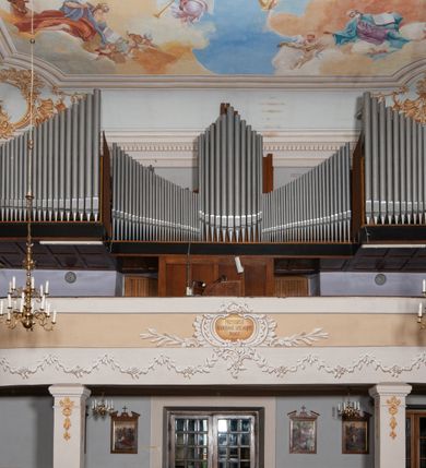 Zdjęcie nr 24: Kościół orientowany, murowany, trójnawowy, z prezbiterium zamkniętym trójbocznie, zakrystią od północy i kruchtą od południa. Do naw przylegają kaplice: większa od północy i mniejsza od południa. Dwie kolejne kaplice przylegają do boków wieży. Ściany wnętrza gładkie, pokryte polichromią imitującą podziały i obramienia, dekorację ornamentalną i sceny iluzjonistyczne na stropach, utrzymane w chłodnej biało-niebiesko-różowej tonacji z dodatkiem żółtego. Na stropie prezbiterium scena Zwiastowania, na stropie nawy Zesłania Ducha Świętego. Od zachodu chór muzyczny wsparty na dwóch filarach z balustradą pokrytą polichromią. Prezbiterium i nawa główna nakryte stropami, nad prezbiterium o przekroju spłaszczonej kolebki, nad nawą płaski, z szerokimi fasetami, zakrystia sklepiona kolebkowo, kruchta i kaplice wschodnie krzyżowo. Nawę, prezbiterium i część kaplic doświetlają okna w kształcie stojącego prostokąta zamkniętego łukiem pełnym, pozostałe zamknięte łukiem odcinkowym lub kwadratowe. Główne wejście i dwa boczne od zachodu, kolejne do przedsionka od północy i do kruchty od południa. Fasada z wieżą na osi, poprzedzona portykiem wspartym na filarach, z trójkątnym szczytem zwieńczonym krzyżem łacińskim. Fasada jednokondygnacyjna, przechodząca w masyw wieży. Kondygnację przepruwają trzy okna i jest zamknięta gzymsem wyniesionym na środku w łuk. Wieńczący ją trójkątny szczyt wtapia się w kondygnację wieży o gładkich elewacjach przeprutych jedynie niewielkimi oknami i tarczami zegarów. Ostatnia kondygnacja jest ujęta gzymsami i dekorowana fryzem arkadowym, którego arkady wykonano z nietynkowanej cegły. Zwieńczenie rozbudowane składa się z iglicy i ustawionych wokół niej, w narożach małych, dwukondygnacyjnych wieżyczek zamkniętych dachami stożkowymi. Wieżyczki połączono podwójnymi arkadami, których łuki wykonano z nietynkowanej cegły, tak samo jak górne kondygnacje wieżyczek. Elewacje kościoła gładkie, na niskim, kamiennym cokole, od wschodu opięte szkarpami. Prezbiterium i nawę przekrywają osobne dachy dwuspadowe, na kalenicy nawy wieżyczka na sygnaturkę, cztery kaplice i portyk nakrywają dachy trójpołaciowe, zakrystię i nawy boczne pulpitowe. 
Jedyne zachowane elementy wyposażenia sprzed pożaru w 1815 roku to fragment osiemnastowiecznej polichromii przedstawiającej dwóch aniołów z narzędziami męki i przedmioty nawiązujące do tego wydarzenia oraz gotycki portal do zakrystii wraz z żelaznymi, kutymi drzwiami. 
