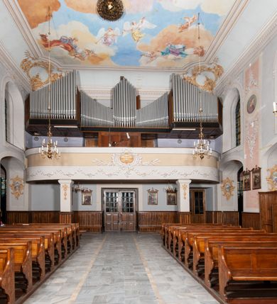 Zdjęcie nr 20: Kościół orientowany, murowany, trójnawowy, z prezbiterium zamkniętym trójbocznie, zakrystią od północy i kruchtą od południa. Do naw przylegają kaplice: większa od północy i mniejsza od południa. Dwie kolejne kaplice przylegają do boków wieży. Ściany wnętrza gładkie, pokryte polichromią imitującą podziały i obramienia, dekorację ornamentalną i sceny iluzjonistyczne na stropach, utrzymane w chłodnej biało-niebiesko-różowej tonacji z dodatkiem żółtego. Na stropie prezbiterium scena Zwiastowania, na stropie nawy Zesłania Ducha Świętego. Od zachodu chór muzyczny wsparty na dwóch filarach z balustradą pokrytą polichromią. Prezbiterium i nawa główna nakryte stropami, nad prezbiterium o przekroju spłaszczonej kolebki, nad nawą płaski, z szerokimi fasetami, zakrystia sklepiona kolebkowo, kruchta i kaplice wschodnie krzyżowo. Nawę, prezbiterium i część kaplic doświetlają okna w kształcie stojącego prostokąta zamkniętego łukiem pełnym, pozostałe zamknięte łukiem odcinkowym lub kwadratowe. Główne wejście i dwa boczne od zachodu, kolejne do przedsionka od północy i do kruchty od południa. Fasada z wieżą na osi, poprzedzona portykiem wspartym na filarach, z trójkątnym szczytem zwieńczonym krzyżem łacińskim. Fasada jednokondygnacyjna, przechodząca w masyw wieży. Kondygnację przepruwają trzy okna i jest zamknięta gzymsem wyniesionym na środku w łuk. Wieńczący ją trójkątny szczyt wtapia się w kondygnację wieży o gładkich elewacjach przeprutych jedynie niewielkimi oknami i tarczami zegarów. Ostatnia kondygnacja jest ujęta gzymsami i dekorowana fryzem arkadowym, którego arkady wykonano z nietynkowanej cegły. Zwieńczenie rozbudowane składa się z iglicy i ustawionych wokół niej, w narożach małych, dwukondygnacyjnych wieżyczek zamkniętych dachami stożkowymi. Wieżyczki połączono podwójnymi arkadami, których łuki wykonano z nietynkowanej cegły, tak samo jak górne kondygnacje wieżyczek. Elewacje kościoła gładkie, na niskim, kamiennym cokole, od wschodu opięte szkarpami. Prezbiterium i nawę przekrywają osobne dachy dwuspadowe, na kalenicy nawy wieżyczka na sygnaturkę, cztery kaplice i portyk nakrywają dachy trójpołaciowe, zakrystię i nawy boczne pulpitowe. 
Jedyne zachowane elementy wyposażenia sprzed pożaru w 1815 roku to fragment osiemnastowiecznej polichromii przedstawiającej dwóch aniołów z narzędziami męki i przedmioty nawiązujące do tego wydarzenia oraz gotycki portal do zakrystii wraz z żelaznymi, kutymi drzwiami. 
