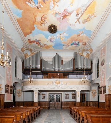 Zdjęcie nr 21: Kościół orientowany, murowany, trójnawowy, z prezbiterium zamkniętym trójbocznie, zakrystią od północy i kruchtą od południa. Do naw przylegają kaplice: większa od północy i mniejsza od południa. Dwie kolejne kaplice przylegają do boków wieży. Ściany wnętrza gładkie, pokryte polichromią imitującą podziały i obramienia, dekorację ornamentalną i sceny iluzjonistyczne na stropach, utrzymane w chłodnej biało-niebiesko-różowej tonacji z dodatkiem żółtego. Na stropie prezbiterium scena Zwiastowania, na stropie nawy Zesłania Ducha Świętego. Od zachodu chór muzyczny wsparty na dwóch filarach z balustradą pokrytą polichromią. Prezbiterium i nawa główna nakryte stropami, nad prezbiterium o przekroju spłaszczonej kolebki, nad nawą płaski, z szerokimi fasetami, zakrystia sklepiona kolebkowo, kruchta i kaplice wschodnie krzyżowo. Nawę, prezbiterium i część kaplic doświetlają okna w kształcie stojącego prostokąta zamkniętego łukiem pełnym, pozostałe zamknięte łukiem odcinkowym lub kwadratowe. Główne wejście i dwa boczne od zachodu, kolejne do przedsionka od północy i do kruchty od południa. Fasada z wieżą na osi, poprzedzona portykiem wspartym na filarach, z trójkątnym szczytem zwieńczonym krzyżem łacińskim. Fasada jednokondygnacyjna, przechodząca w masyw wieży. Kondygnację przepruwają trzy okna i jest zamknięta gzymsem wyniesionym na środku w łuk. Wieńczący ją trójkątny szczyt wtapia się w kondygnację wieży o gładkich elewacjach przeprutych jedynie niewielkimi oknami i tarczami zegarów. Ostatnia kondygnacja jest ujęta gzymsami i dekorowana fryzem arkadowym, którego arkady wykonano z nietynkowanej cegły. Zwieńczenie rozbudowane składa się z iglicy i ustawionych wokół niej, w narożach małych, dwukondygnacyjnych wieżyczek zamkniętych dachami stożkowymi. Wieżyczki połączono podwójnymi arkadami, których łuki wykonano z nietynkowanej cegły, tak samo jak górne kondygnacje wieżyczek. Elewacje kościoła gładkie, na niskim, kamiennym cokole, od wschodu opięte szkarpami. Prezbiterium i nawę przekrywają osobne dachy dwuspadowe, na kalenicy nawy wieżyczka na sygnaturkę, cztery kaplice i portyk nakrywają dachy trójpołaciowe, zakrystię i nawy boczne pulpitowe. 
Jedyne zachowane elementy wyposażenia sprzed pożaru w 1815 roku to fragment osiemnastowiecznej polichromii przedstawiającej dwóch aniołów z narzędziami męki i przedmioty nawiązujące do tego wydarzenia oraz gotycki portal do zakrystii wraz z żelaznymi, kutymi drzwiami. 
