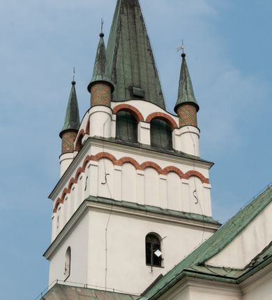 Zdjęcie nr 11: Kościół orientowany, murowany, trójnawowy, z prezbiterium zamkniętym trójbocznie, zakrystią od północy i kruchtą od południa. Do naw przylegają kaplice: większa od północy i mniejsza od południa. Dwie kolejne kaplice przylegają do boków wieży. Ściany wnętrza gładkie, pokryte polichromią imitującą podziały i obramienia, dekorację ornamentalną i sceny iluzjonistyczne na stropach, utrzymane w chłodnej biało-niebiesko-różowej tonacji z dodatkiem żółtego. Na stropie prezbiterium scena Zwiastowania, na stropie nawy Zesłania Ducha Świętego. Od zachodu chór muzyczny wsparty na dwóch filarach z balustradą pokrytą polichromią. Prezbiterium i nawa główna nakryte stropami, nad prezbiterium o przekroju spłaszczonej kolebki, nad nawą płaski, z szerokimi fasetami, zakrystia sklepiona kolebkowo, kruchta i kaplice wschodnie krzyżowo. Nawę, prezbiterium i część kaplic doświetlają okna w kształcie stojącego prostokąta zamkniętego łukiem pełnym, pozostałe zamknięte łukiem odcinkowym lub kwadratowe. Główne wejście i dwa boczne od zachodu, kolejne do przedsionka od północy i do kruchty od południa. Fasada z wieżą na osi, poprzedzona portykiem wspartym na filarach, z trójkątnym szczytem zwieńczonym krzyżem łacińskim. Fasada jednokondygnacyjna, przechodząca w masyw wieży. Kondygnację przepruwają trzy okna i jest zamknięta gzymsem wyniesionym na środku w łuk. Wieńczący ją trójkątny szczyt wtapia się w kondygnację wieży o gładkich elewacjach przeprutych jedynie niewielkimi oknami i tarczami zegarów. Ostatnia kondygnacja jest ujęta gzymsami i dekorowana fryzem arkadowym, którego arkady wykonano z nietynkowanej cegły. Zwieńczenie rozbudowane składa się z iglicy i ustawionych wokół niej, w narożach małych, dwukondygnacyjnych wieżyczek zamkniętych dachami stożkowymi. Wieżyczki połączono podwójnymi arkadami, których łuki wykonano z nietynkowanej cegły, tak samo jak górne kondygnacje wieżyczek. Elewacje kościoła gładkie, na niskim, kamiennym cokole, od wschodu opięte szkarpami. Prezbiterium i nawę przekrywają osobne dachy dwuspadowe, na kalenicy nawy wieżyczka na sygnaturkę, cztery kaplice i portyk nakrywają dachy trójpołaciowe, zakrystię i nawy boczne pulpitowe. 
Jedyne zachowane elementy wyposażenia sprzed pożaru w 1815 roku to fragment osiemnastowiecznej polichromii przedstawiającej dwóch aniołów z narzędziami męki i przedmioty nawiązujące do tego wydarzenia oraz gotycki portal do zakrystii wraz z żelaznymi, kutymi drzwiami. 
