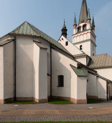 Zdjęcie nr 10: Kościół orientowany, murowany, trójnawowy, z prezbiterium zamkniętym trójbocznie, zakrystią od północy i kruchtą od południa. Do naw przylegają kaplice: większa od północy i mniejsza od południa. Dwie kolejne kaplice przylegają do boków wieży. Ściany wnętrza gładkie, pokryte polichromią imitującą podziały i obramienia, dekorację ornamentalną i sceny iluzjonistyczne na stropach, utrzymane w chłodnej biało-niebiesko-różowej tonacji z dodatkiem żółtego. Na stropie prezbiterium scena Zwiastowania, na stropie nawy Zesłania Ducha Świętego. Od zachodu chór muzyczny wsparty na dwóch filarach z balustradą pokrytą polichromią. Prezbiterium i nawa główna nakryte stropami, nad prezbiterium o przekroju spłaszczonej kolebki, nad nawą płaski, z szerokimi fasetami, zakrystia sklepiona kolebkowo, kruchta i kaplice wschodnie krzyżowo. Nawę, prezbiterium i część kaplic doświetlają okna w kształcie stojącego prostokąta zamkniętego łukiem pełnym, pozostałe zamknięte łukiem odcinkowym lub kwadratowe. Główne wejście i dwa boczne od zachodu, kolejne do przedsionka od północy i do kruchty od południa. Fasada z wieżą na osi, poprzedzona portykiem wspartym na filarach, z trójkątnym szczytem zwieńczonym krzyżem łacińskim. Fasada jednokondygnacyjna, przechodząca w masyw wieży. Kondygnację przepruwają trzy okna i jest zamknięta gzymsem wyniesionym na środku w łuk. Wieńczący ją trójkątny szczyt wtapia się w kondygnację wieży o gładkich elewacjach przeprutych jedynie niewielkimi oknami i tarczami zegarów. Ostatnia kondygnacja jest ujęta gzymsami i dekorowana fryzem arkadowym, którego arkady wykonano z nietynkowanej cegły. Zwieńczenie rozbudowane składa się z iglicy i ustawionych wokół niej, w narożach małych, dwukondygnacyjnych wieżyczek zamkniętych dachami stożkowymi. Wieżyczki połączono podwójnymi arkadami, których łuki wykonano z nietynkowanej cegły, tak samo jak górne kondygnacje wieżyczek. Elewacje kościoła gładkie, na niskim, kamiennym cokole, od wschodu opięte szkarpami. Prezbiterium i nawę przekrywają osobne dachy dwuspadowe, na kalenicy nawy wieżyczka na sygnaturkę, cztery kaplice i portyk nakrywają dachy trójpołaciowe, zakrystię i nawy boczne pulpitowe. 
Jedyne zachowane elementy wyposażenia sprzed pożaru w 1815 roku to fragment osiemnastowiecznej polichromii przedstawiającej dwóch aniołów z narzędziami męki i przedmioty nawiązujące do tego wydarzenia oraz gotycki portal do zakrystii wraz z żelaznymi, kutymi drzwiami. 
