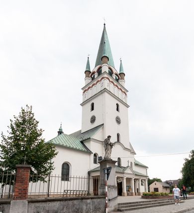 Zdjęcie nr 2: Kościół orientowany, murowany, trójnawowy, z prezbiterium zamkniętym trójbocznie, zakrystią od północy i kruchtą od południa. Do naw przylegają kaplice: większa od północy i mniejsza od południa. Dwie kolejne kaplice przylegają do boków wieży. Ściany wnętrza gładkie, pokryte polichromią imitującą podziały i obramienia, dekorację ornamentalną i sceny iluzjonistyczne na stropach, utrzymane w chłodnej biało-niebiesko-różowej tonacji z dodatkiem żółtego. Na stropie prezbiterium scena Zwiastowania, na stropie nawy Zesłania Ducha Świętego. Od zachodu chór muzyczny wsparty na dwóch filarach z balustradą pokrytą polichromią. Prezbiterium i nawa główna nakryte stropami, nad prezbiterium o przekroju spłaszczonej kolebki, nad nawą płaski, z szerokimi fasetami, zakrystia sklepiona kolebkowo, kruchta i kaplice wschodnie krzyżowo. Nawę, prezbiterium i część kaplic doświetlają okna w kształcie stojącego prostokąta zamkniętego łukiem pełnym, pozostałe zamknięte łukiem odcinkowym lub kwadratowe. Główne wejście i dwa boczne od zachodu, kolejne do przedsionka od północy i do kruchty od południa. Fasada z wieżą na osi, poprzedzona portykiem wspartym na filarach, z trójkątnym szczytem zwieńczonym krzyżem łacińskim. Fasada jednokondygnacyjna, przechodząca w masyw wieży. Kondygnację przepruwają trzy okna i jest zamknięta gzymsem wyniesionym na środku w łuk. Wieńczący ją trójkątny szczyt wtapia się w kondygnację wieży o gładkich elewacjach przeprutych jedynie niewielkimi oknami i tarczami zegarów. Ostatnia kondygnacja jest ujęta gzymsami i dekorowana fryzem arkadowym, którego arkady wykonano z nietynkowanej cegły. Zwieńczenie rozbudowane składa się z iglicy i ustawionych wokół niej, w narożach małych, dwukondygnacyjnych wieżyczek zamkniętych dachami stożkowymi. Wieżyczki połączono podwójnymi arkadami, których łuki wykonano z nietynkowanej cegły, tak samo jak górne kondygnacje wieżyczek. Elewacje kościoła gładkie, na niskim, kamiennym cokole, od wschodu opięte szkarpami. Prezbiterium i nawę przekrywają osobne dachy dwuspadowe, na kalenicy nawy wieżyczka na sygnaturkę, cztery kaplice i portyk nakrywają dachy trójpołaciowe, zakrystię i nawy boczne pulpitowe. 
Jedyne zachowane elementy wyposażenia sprzed pożaru w 1815 roku to fragment osiemnastowiecznej polichromii przedstawiającej dwóch aniołów z narzędziami męki i przedmioty nawiązujące do tego wydarzenia oraz gotycki portal do zakrystii wraz z żelaznymi, kutymi drzwiami. 
