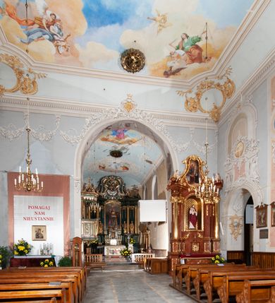 Zdjęcie nr 13: Kościół orientowany, murowany, trójnawowy, z prezbiterium zamkniętym trójbocznie, zakrystią od północy i kruchtą od południa. Do naw przylegają kaplice: większa od północy i mniejsza od południa. Dwie kolejne kaplice przylegają do boków wieży. Ściany wnętrza gładkie, pokryte polichromią imitującą podziały i obramienia, dekorację ornamentalną i sceny iluzjonistyczne na stropach, utrzymane w chłodnej biało-niebiesko-różowej tonacji z dodatkiem żółtego. Na stropie prezbiterium scena Zwiastowania, na stropie nawy Zesłania Ducha Świętego. Od zachodu chór muzyczny wsparty na dwóch filarach z balustradą pokrytą polichromią. Prezbiterium i nawa główna nakryte stropami, nad prezbiterium o przekroju spłaszczonej kolebki, nad nawą płaski, z szerokimi fasetami, zakrystia sklepiona kolebkowo, kruchta i kaplice wschodnie krzyżowo. Nawę, prezbiterium i część kaplic doświetlają okna w kształcie stojącego prostokąta zamkniętego łukiem pełnym, pozostałe zamknięte łukiem odcinkowym lub kwadratowe. Główne wejście i dwa boczne od zachodu, kolejne do przedsionka od północy i do kruchty od południa. Fasada z wieżą na osi, poprzedzona portykiem wspartym na filarach, z trójkątnym szczytem zwieńczonym krzyżem łacińskim. Fasada jednokondygnacyjna, przechodząca w masyw wieży. Kondygnację przepruwają trzy okna i jest zamknięta gzymsem wyniesionym na środku w łuk. Wieńczący ją trójkątny szczyt wtapia się w kondygnację wieży o gładkich elewacjach przeprutych jedynie niewielkimi oknami i tarczami zegarów. Ostatnia kondygnacja jest ujęta gzymsami i dekorowana fryzem arkadowym, którego arkady wykonano z nietynkowanej cegły. Zwieńczenie rozbudowane składa się z iglicy i ustawionych wokół niej, w narożach małych, dwukondygnacyjnych wieżyczek zamkniętych dachami stożkowymi. Wieżyczki połączono podwójnymi arkadami, których łuki wykonano z nietynkowanej cegły, tak samo jak górne kondygnacje wieżyczek. Elewacje kościoła gładkie, na niskim, kamiennym cokole, od wschodu opięte szkarpami. Prezbiterium i nawę przekrywają osobne dachy dwuspadowe, na kalenicy nawy wieżyczka na sygnaturkę, cztery kaplice i portyk nakrywają dachy trójpołaciowe, zakrystię i nawy boczne pulpitowe. 
Jedyne zachowane elementy wyposażenia sprzed pożaru w 1815 roku to fragment osiemnastowiecznej polichromii przedstawiającej dwóch aniołów z narzędziami męki i przedmioty nawiązujące do tego wydarzenia oraz gotycki portal do zakrystii wraz z żelaznymi, kutymi drzwiami. 
