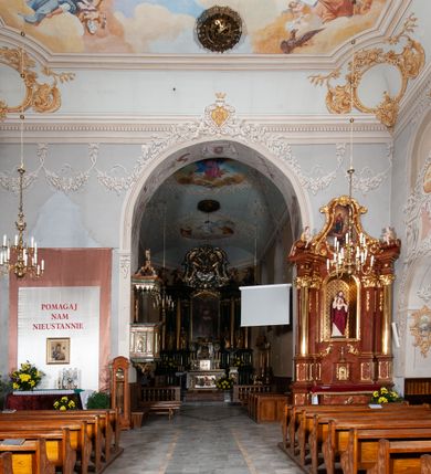 Zdjęcie nr 16: Kościół orientowany, murowany, trójnawowy, z prezbiterium zamkniętym trójbocznie, zakrystią od północy i kruchtą od południa. Do naw przylegają kaplice: większa od północy i mniejsza od południa. Dwie kolejne kaplice przylegają do boków wieży. Ściany wnętrza gładkie, pokryte polichromią imitującą podziały i obramienia, dekorację ornamentalną i sceny iluzjonistyczne na stropach, utrzymane w chłodnej biało-niebiesko-różowej tonacji z dodatkiem żółtego. Na stropie prezbiterium scena Zwiastowania, na stropie nawy Zesłania Ducha Świętego. Od zachodu chór muzyczny wsparty na dwóch filarach z balustradą pokrytą polichromią. Prezbiterium i nawa główna nakryte stropami, nad prezbiterium o przekroju spłaszczonej kolebki, nad nawą płaski, z szerokimi fasetami, zakrystia sklepiona kolebkowo, kruchta i kaplice wschodnie krzyżowo. Nawę, prezbiterium i część kaplic doświetlają okna w kształcie stojącego prostokąta zamkniętego łukiem pełnym, pozostałe zamknięte łukiem odcinkowym lub kwadratowe. Główne wejście i dwa boczne od zachodu, kolejne do przedsionka od północy i do kruchty od południa. Fasada z wieżą na osi, poprzedzona portykiem wspartym na filarach, z trójkątnym szczytem zwieńczonym krzyżem łacińskim. Fasada jednokondygnacyjna, przechodząca w masyw wieży. Kondygnację przepruwają trzy okna i jest zamknięta gzymsem wyniesionym na środku w łuk. Wieńczący ją trójkątny szczyt wtapia się w kondygnację wieży o gładkich elewacjach przeprutych jedynie niewielkimi oknami i tarczami zegarów. Ostatnia kondygnacja jest ujęta gzymsami i dekorowana fryzem arkadowym, którego arkady wykonano z nietynkowanej cegły. Zwieńczenie rozbudowane składa się z iglicy i ustawionych wokół niej, w narożach małych, dwukondygnacyjnych wieżyczek zamkniętych dachami stożkowymi. Wieżyczki połączono podwójnymi arkadami, których łuki wykonano z nietynkowanej cegły, tak samo jak górne kondygnacje wieżyczek. Elewacje kościoła gładkie, na niskim, kamiennym cokole, od wschodu opięte szkarpami. Prezbiterium i nawę przekrywają osobne dachy dwuspadowe, na kalenicy nawy wieżyczka na sygnaturkę, cztery kaplice i portyk nakrywają dachy trójpołaciowe, zakrystię i nawy boczne pulpitowe. 
Jedyne zachowane elementy wyposażenia sprzed pożaru w 1815 roku to fragment osiemnastowiecznej polichromii przedstawiającej dwóch aniołów z narzędziami męki i przedmioty nawiązujące do tego wydarzenia oraz gotycki portal do zakrystii wraz z żelaznymi, kutymi drzwiami. 
