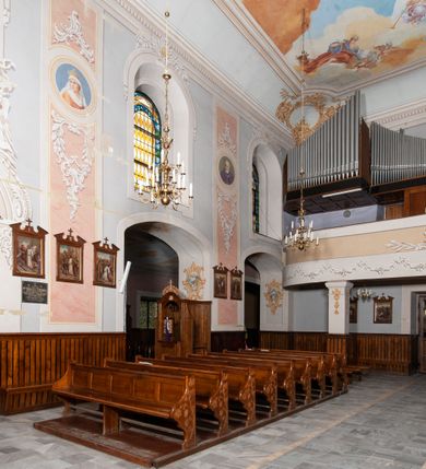 Zdjęcie nr 26: Kościół orientowany, murowany, trójnawowy, z prezbiterium zamkniętym trójbocznie, zakrystią od północy i kruchtą od południa. Do naw przylegają kaplice: większa od północy i mniejsza od południa. Dwie kolejne kaplice przylegają do boków wieży. Ściany wnętrza gładkie, pokryte polichromią imitującą podziały i obramienia, dekorację ornamentalną i sceny iluzjonistyczne na stropach, utrzymane w chłodnej biało-niebiesko-różowej tonacji z dodatkiem żółtego. Na stropie prezbiterium scena Zwiastowania, na stropie nawy Zesłania Ducha Świętego. Od zachodu chór muzyczny wsparty na dwóch filarach z balustradą pokrytą polichromią. Prezbiterium i nawa główna nakryte stropami, nad prezbiterium o przekroju spłaszczonej kolebki, nad nawą płaski, z szerokimi fasetami, zakrystia sklepiona kolebkowo, kruchta i kaplice wschodnie krzyżowo. Nawę, prezbiterium i część kaplic doświetlają okna w kształcie stojącego prostokąta zamkniętego łukiem pełnym, pozostałe zamknięte łukiem odcinkowym lub kwadratowe. Główne wejście i dwa boczne od zachodu, kolejne do przedsionka od północy i do kruchty od południa. Fasada z wieżą na osi, poprzedzona portykiem wspartym na filarach, z trójkątnym szczytem zwieńczonym krzyżem łacińskim. Fasada jednokondygnacyjna, przechodząca w masyw wieży. Kondygnację przepruwają trzy okna i jest zamknięta gzymsem wyniesionym na środku w łuk. Wieńczący ją trójkątny szczyt wtapia się w kondygnację wieży o gładkich elewacjach przeprutych jedynie niewielkimi oknami i tarczami zegarów. Ostatnia kondygnacja jest ujęta gzymsami i dekorowana fryzem arkadowym, którego arkady wykonano z nietynkowanej cegły. Zwieńczenie rozbudowane składa się z iglicy i ustawionych wokół niej, w narożach małych, dwukondygnacyjnych wieżyczek zamkniętych dachami stożkowymi. Wieżyczki połączono podwójnymi arkadami, których łuki wykonano z nietynkowanej cegły, tak samo jak górne kondygnacje wieżyczek. Elewacje kościoła gładkie, na niskim, kamiennym cokole, od wschodu opięte szkarpami. Prezbiterium i nawę przekrywają osobne dachy dwuspadowe, na kalenicy nawy wieżyczka na sygnaturkę, cztery kaplice i portyk nakrywają dachy trójpołaciowe, zakrystię i nawy boczne pulpitowe. 
Jedyne zachowane elementy wyposażenia sprzed pożaru w 1815 roku to fragment osiemnastowiecznej polichromii przedstawiającej dwóch aniołów z narzędziami męki i przedmioty nawiązujące do tego wydarzenia oraz gotycki portal do zakrystii wraz z żelaznymi, kutymi drzwiami. 
