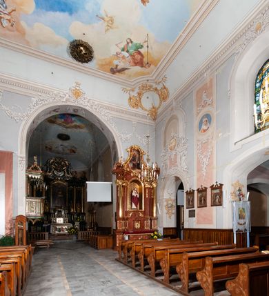 Zdjęcie nr 17: Kościół orientowany, murowany, trójnawowy, z prezbiterium zamkniętym trójbocznie, zakrystią od północy i kruchtą od południa. Do naw przylegają kaplice: większa od północy i mniejsza od południa. Dwie kolejne kaplice przylegają do boków wieży. Ściany wnętrza gładkie, pokryte polichromią imitującą podziały i obramienia, dekorację ornamentalną i sceny iluzjonistyczne na stropach, utrzymane w chłodnej biało-niebiesko-różowej tonacji z dodatkiem żółtego. Na stropie prezbiterium scena Zwiastowania, na stropie nawy Zesłania Ducha Świętego. Od zachodu chór muzyczny wsparty na dwóch filarach z balustradą pokrytą polichromią. Prezbiterium i nawa główna nakryte stropami, nad prezbiterium o przekroju spłaszczonej kolebki, nad nawą płaski, z szerokimi fasetami, zakrystia sklepiona kolebkowo, kruchta i kaplice wschodnie krzyżowo. Nawę, prezbiterium i część kaplic doświetlają okna w kształcie stojącego prostokąta zamkniętego łukiem pełnym, pozostałe zamknięte łukiem odcinkowym lub kwadratowe. Główne wejście i dwa boczne od zachodu, kolejne do przedsionka od północy i do kruchty od południa. Fasada z wieżą na osi, poprzedzona portykiem wspartym na filarach, z trójkątnym szczytem zwieńczonym krzyżem łacińskim. Fasada jednokondygnacyjna, przechodząca w masyw wieży. Kondygnację przepruwają trzy okna i jest zamknięta gzymsem wyniesionym na środku w łuk. Wieńczący ją trójkątny szczyt wtapia się w kondygnację wieży o gładkich elewacjach przeprutych jedynie niewielkimi oknami i tarczami zegarów. Ostatnia kondygnacja jest ujęta gzymsami i dekorowana fryzem arkadowym, którego arkady wykonano z nietynkowanej cegły. Zwieńczenie rozbudowane składa się z iglicy i ustawionych wokół niej, w narożach małych, dwukondygnacyjnych wieżyczek zamkniętych dachami stożkowymi. Wieżyczki połączono podwójnymi arkadami, których łuki wykonano z nietynkowanej cegły, tak samo jak górne kondygnacje wieżyczek. Elewacje kościoła gładkie, na niskim, kamiennym cokole, od wschodu opięte szkarpami. Prezbiterium i nawę przekrywają osobne dachy dwuspadowe, na kalenicy nawy wieżyczka na sygnaturkę, cztery kaplice i portyk nakrywają dachy trójpołaciowe, zakrystię i nawy boczne pulpitowe. 
Jedyne zachowane elementy wyposażenia sprzed pożaru w 1815 roku to fragment osiemnastowiecznej polichromii przedstawiającej dwóch aniołów z narzędziami męki i przedmioty nawiązujące do tego wydarzenia oraz gotycki portal do zakrystii wraz z żelaznymi, kutymi drzwiami. 
